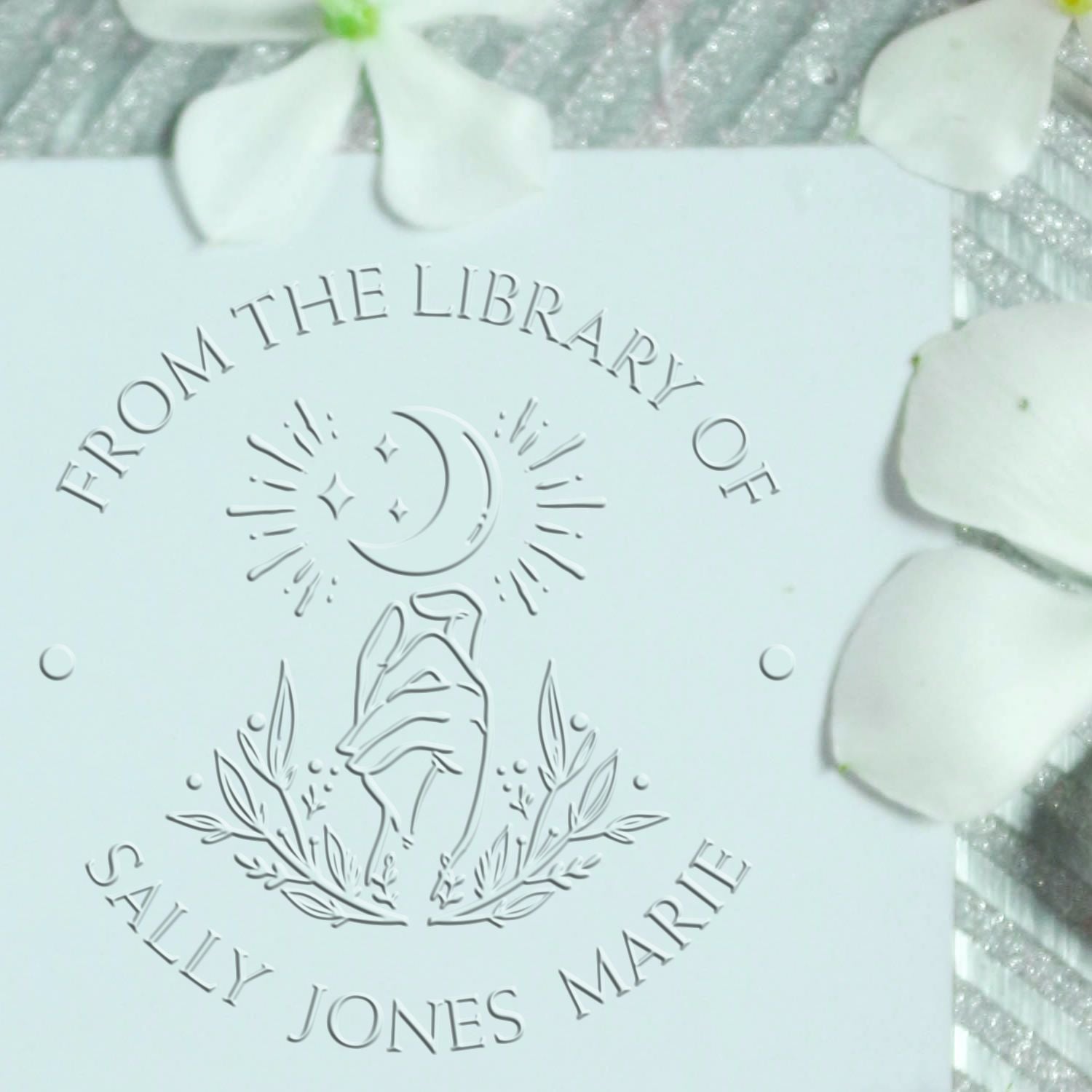White embossed library stamp seal with moon, hand and leaves, reading From the Library of Sally Jones Marie.