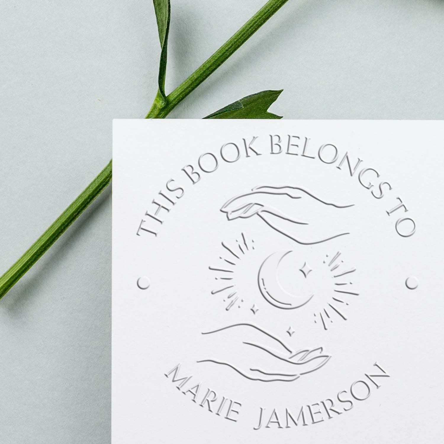White embossed stamp and seal bookplate with hands, moon and stars on pale background and green leaves