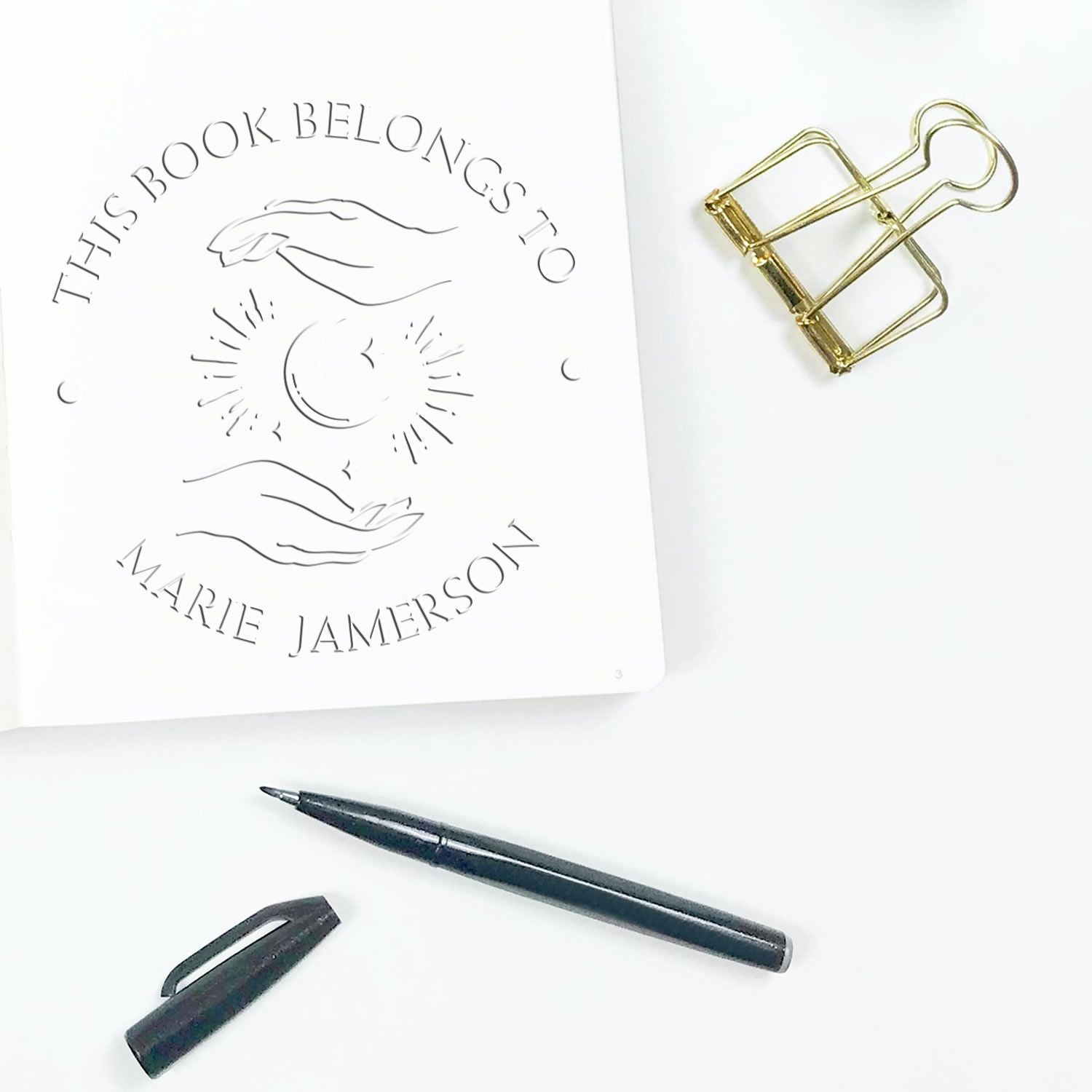 Custom bookplate stamp and seal on white paper, with gold binder clip and black pen and cap.