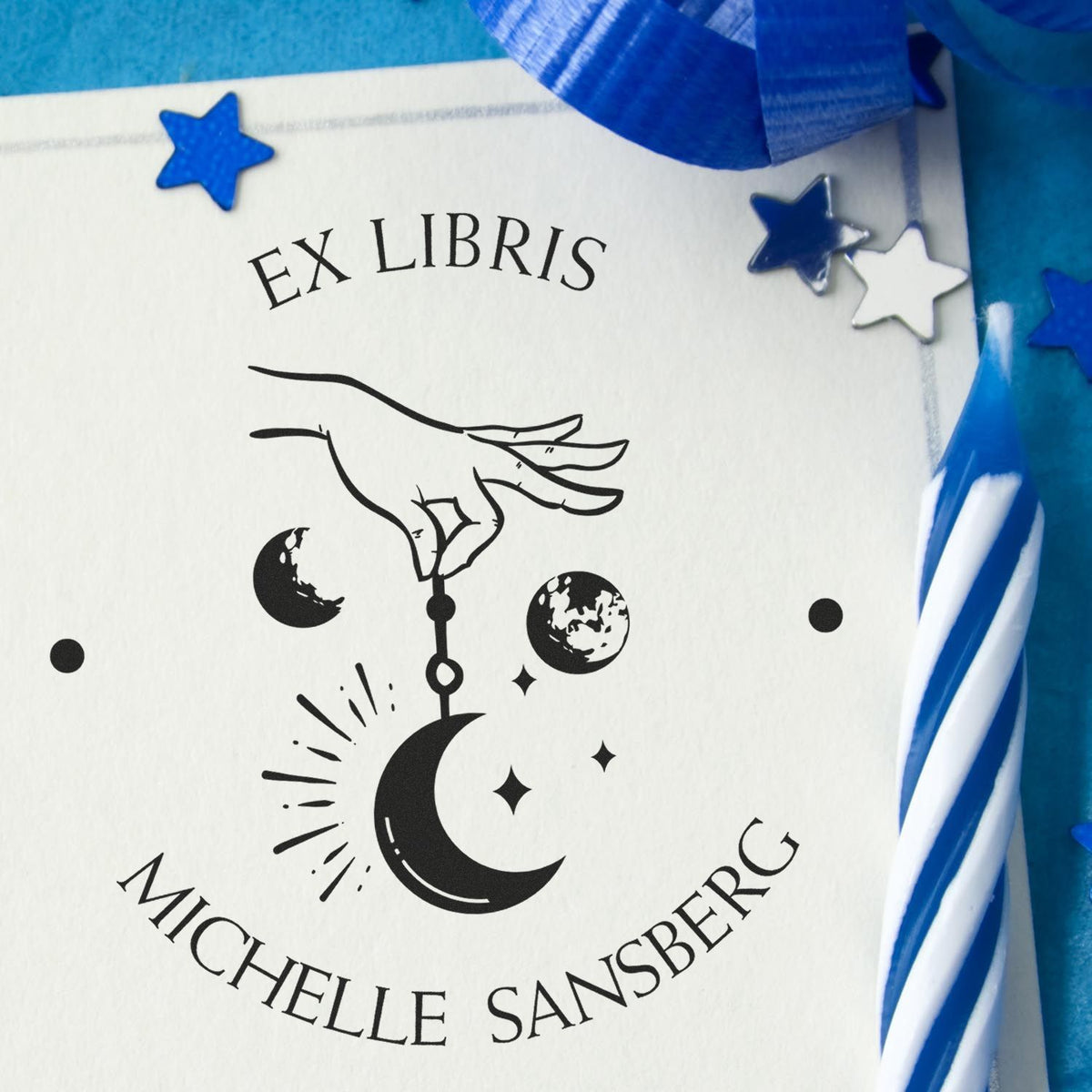 Blue and white Ex Libris stamp seal with hand, crescent moon, stars, and planets, surrounded by blue ribbon and star confetti.