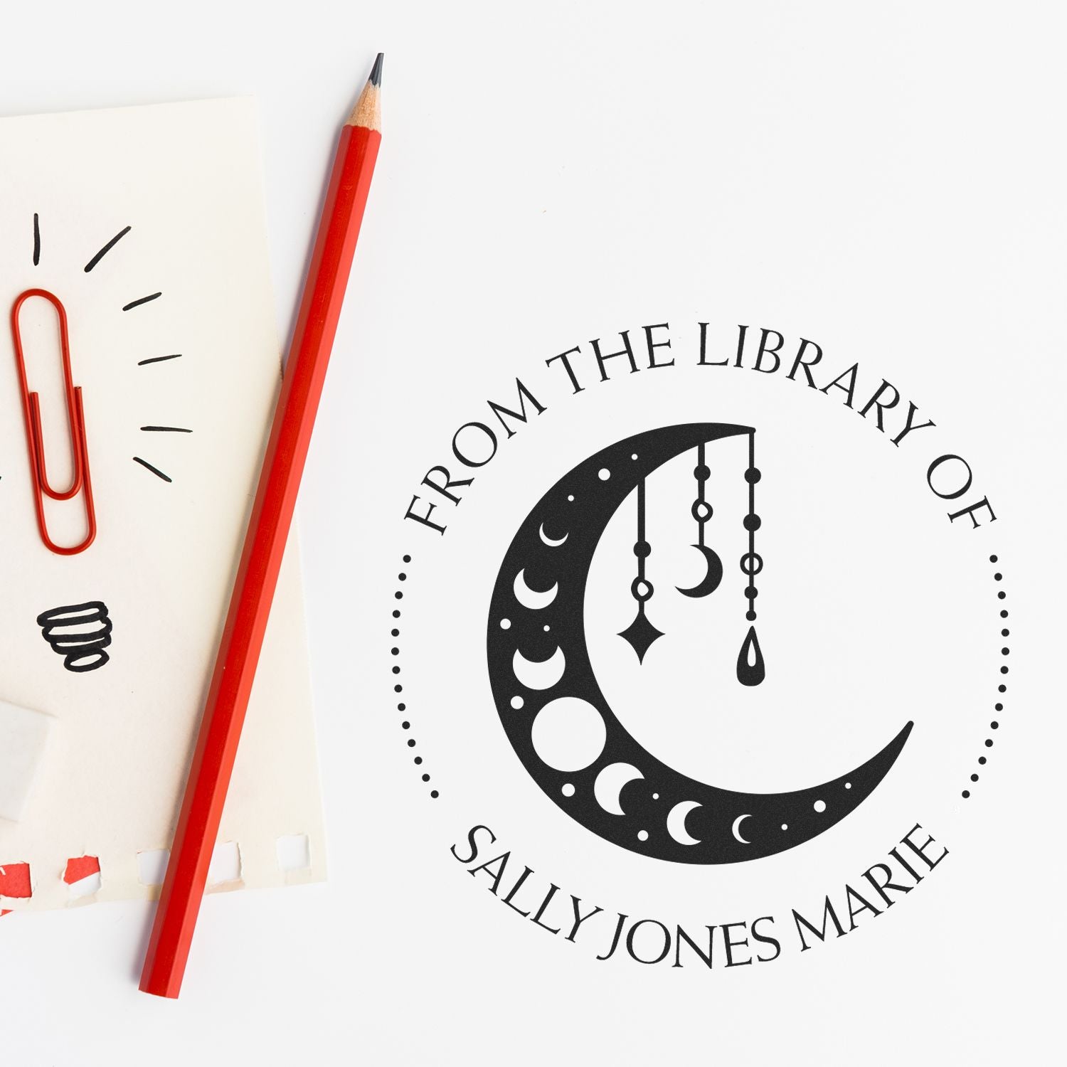 Custom black library stamp seal with crescent moon and hanging charms, beside a red pencil and paperclip on white paper.