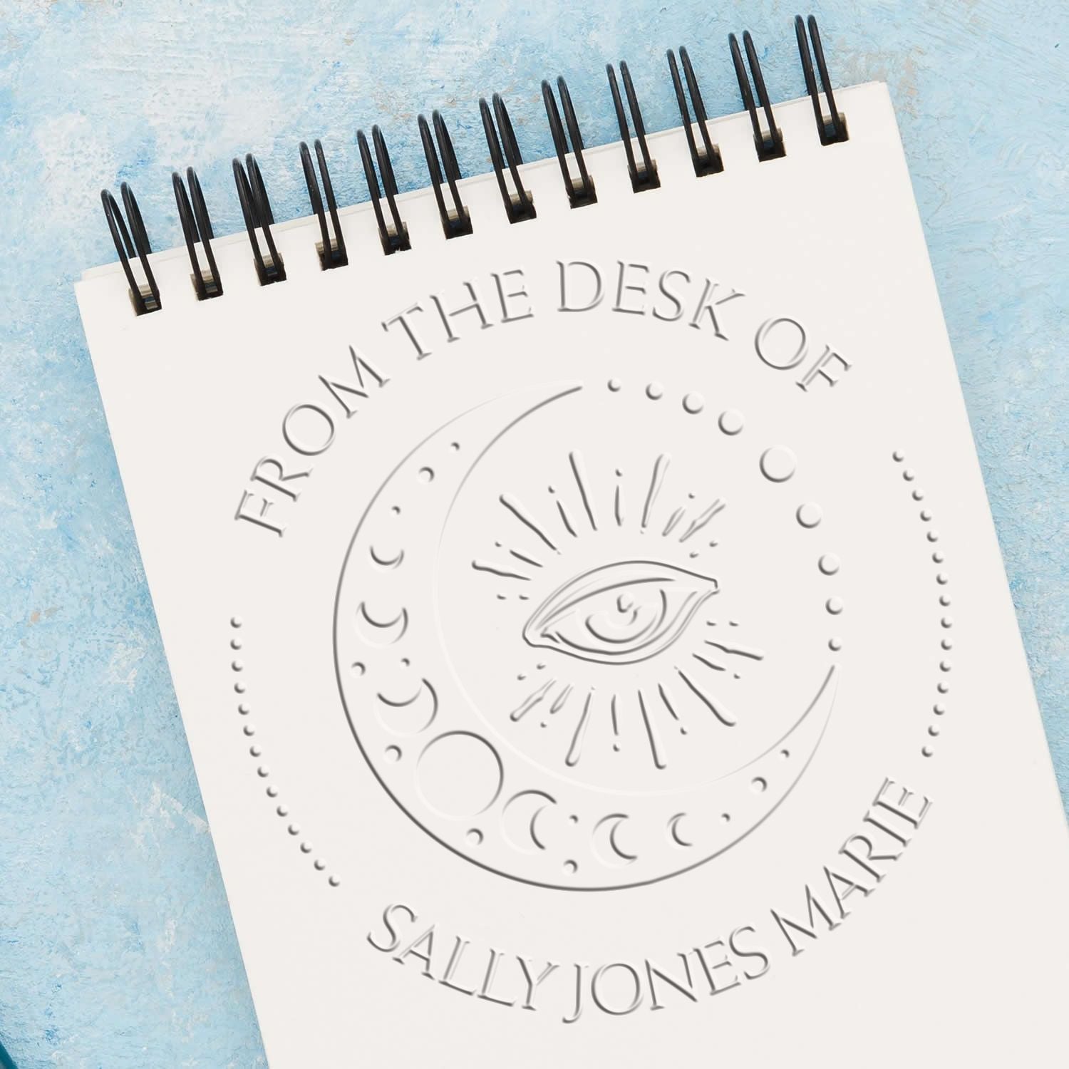 White spiral notepad with embossed moon-eye seal stamp and From the Desk of Sally Jones Marie on a light blue background.