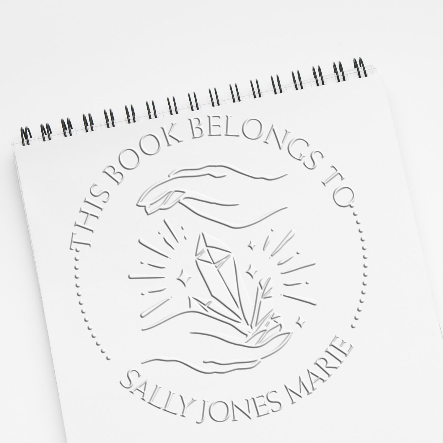 White spiral notebook with embossed stamp-style seal reading This Book Belongs to Sally Jones Marie and crystal hand logo.