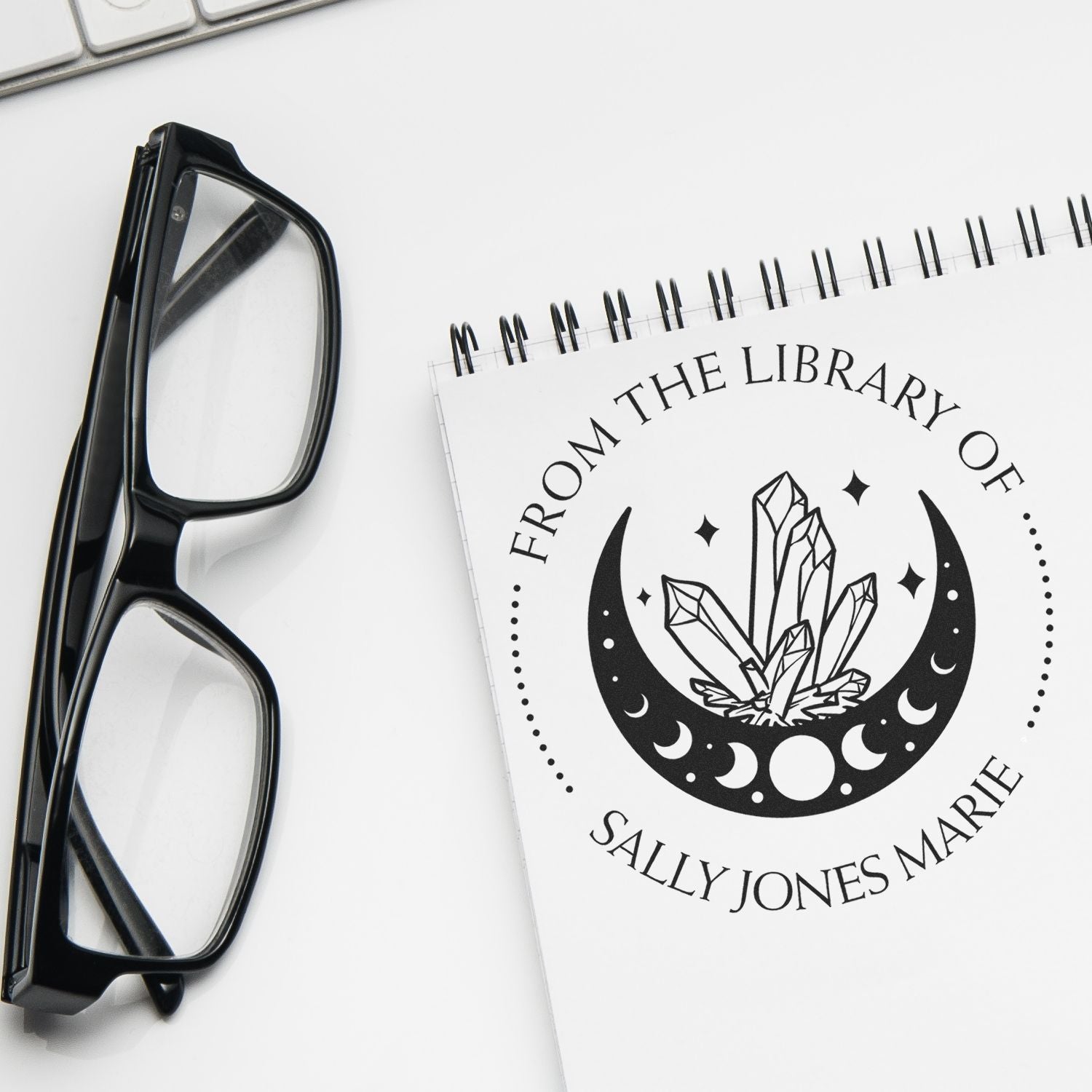 Custom black stamp seal on a white spiral notebook beside black eyeglasses, featuring a crystal moon design.