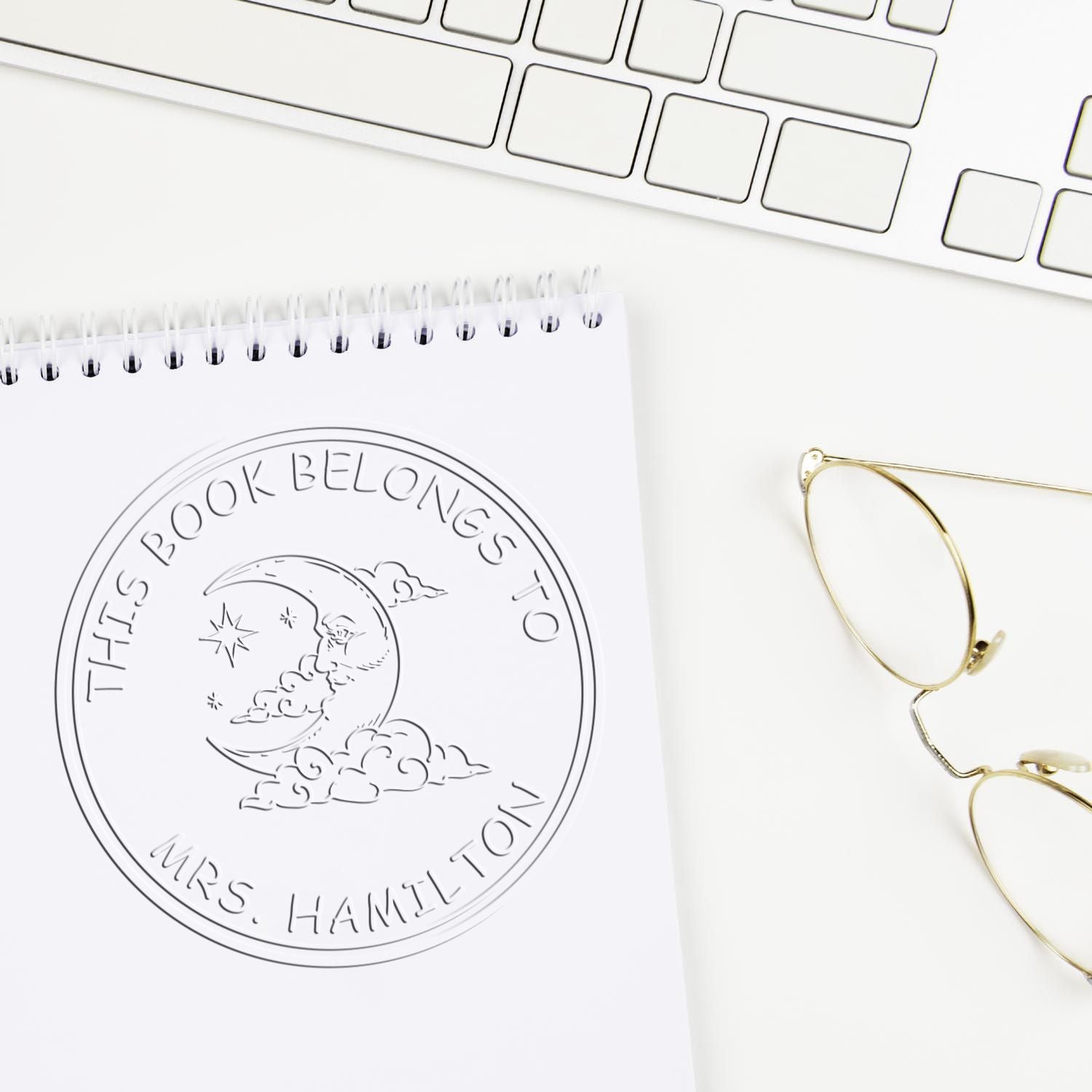 White custom embossed book seal on notepad with moon and clouds design, gold eyeglasses, and keyboard on a white desk.