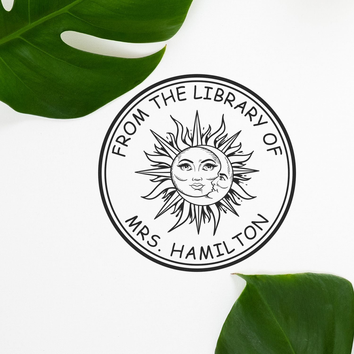 Black custom library stamp seal reading From the Library of Mrs. Hamilton, with sun face design and green leaves on white background