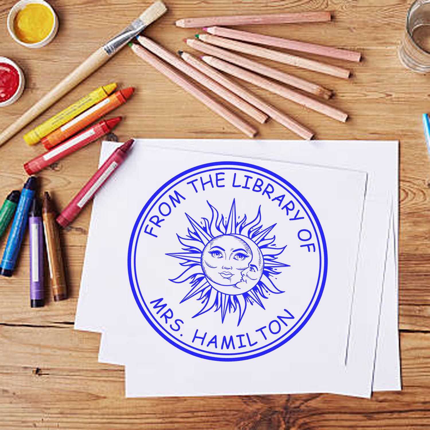 Blue round library seal stamp on white paper, with colored pencils, crayons, paint pots, and brush on a wood table.