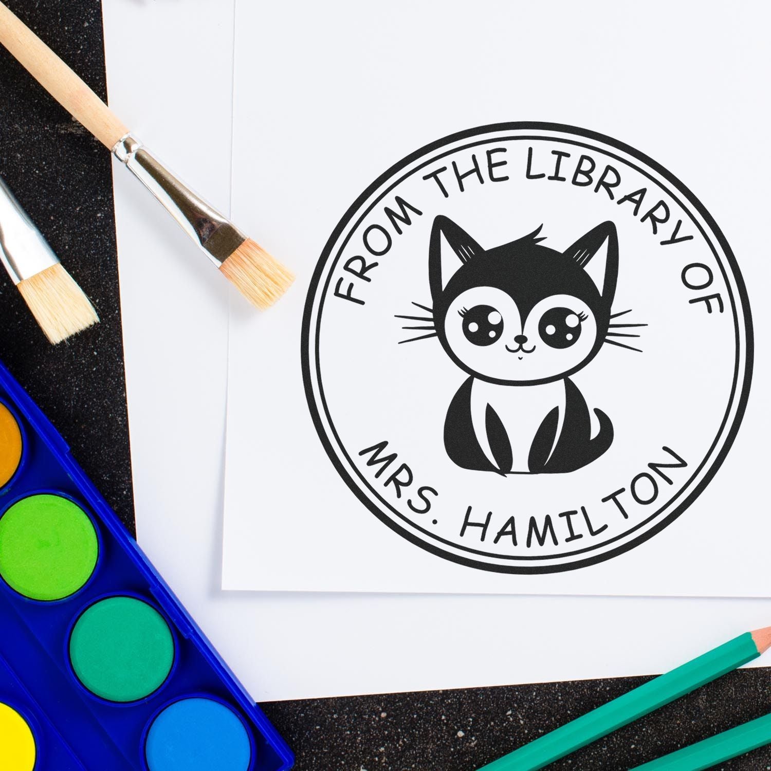 Black custom library stamp seal with cat logo and Mrs. Hamilton text on white paper, surrounded by brushes and paint palette.