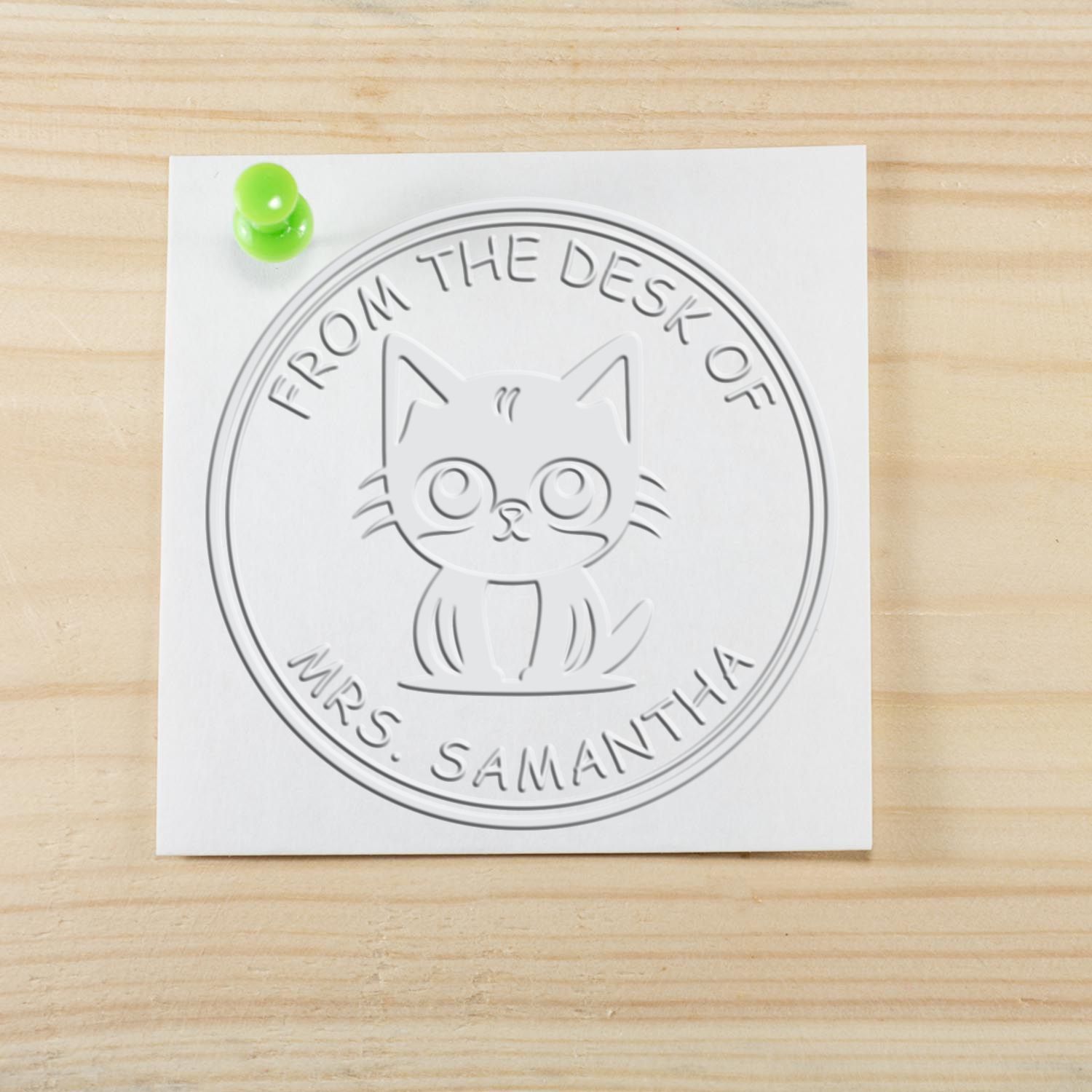 White embossed cat stamp seal on paper, pinned with a green pushpin, on light wood; reads From the Desk of Mrs. Samantha