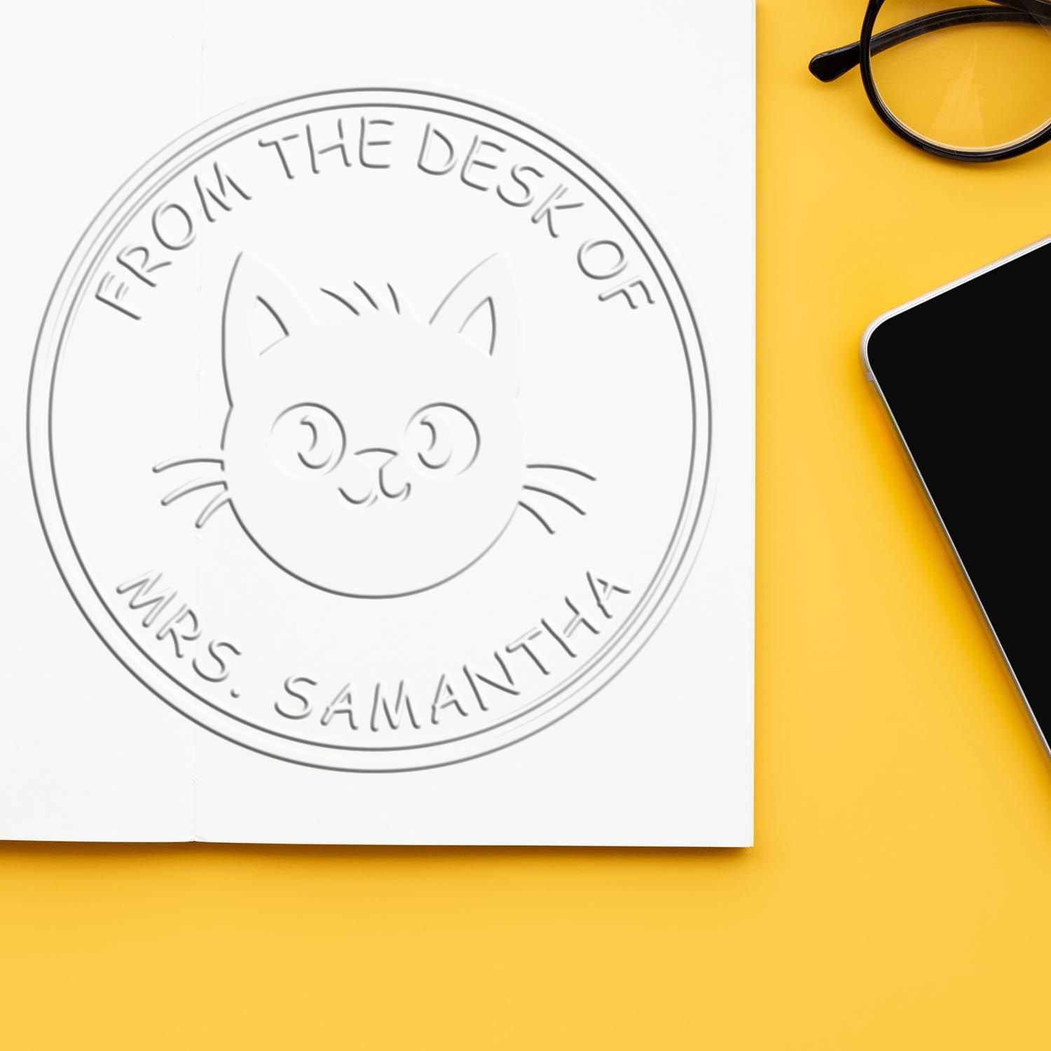 Embossed cat desk seal on white paper, reading From the desk of Mrs. Samantha, with yellow background, glasses and phone.