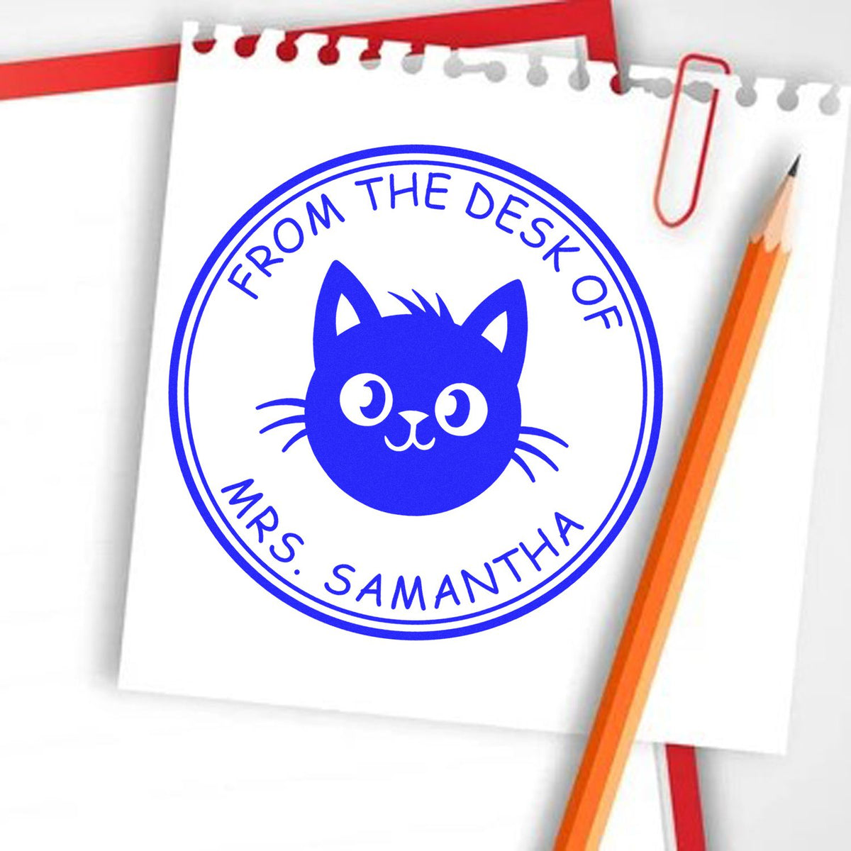 Blue custom desk stamp seal with cat logo and From the desk of Mrs. Samantha on white paper, beside red paperclip and pencil.