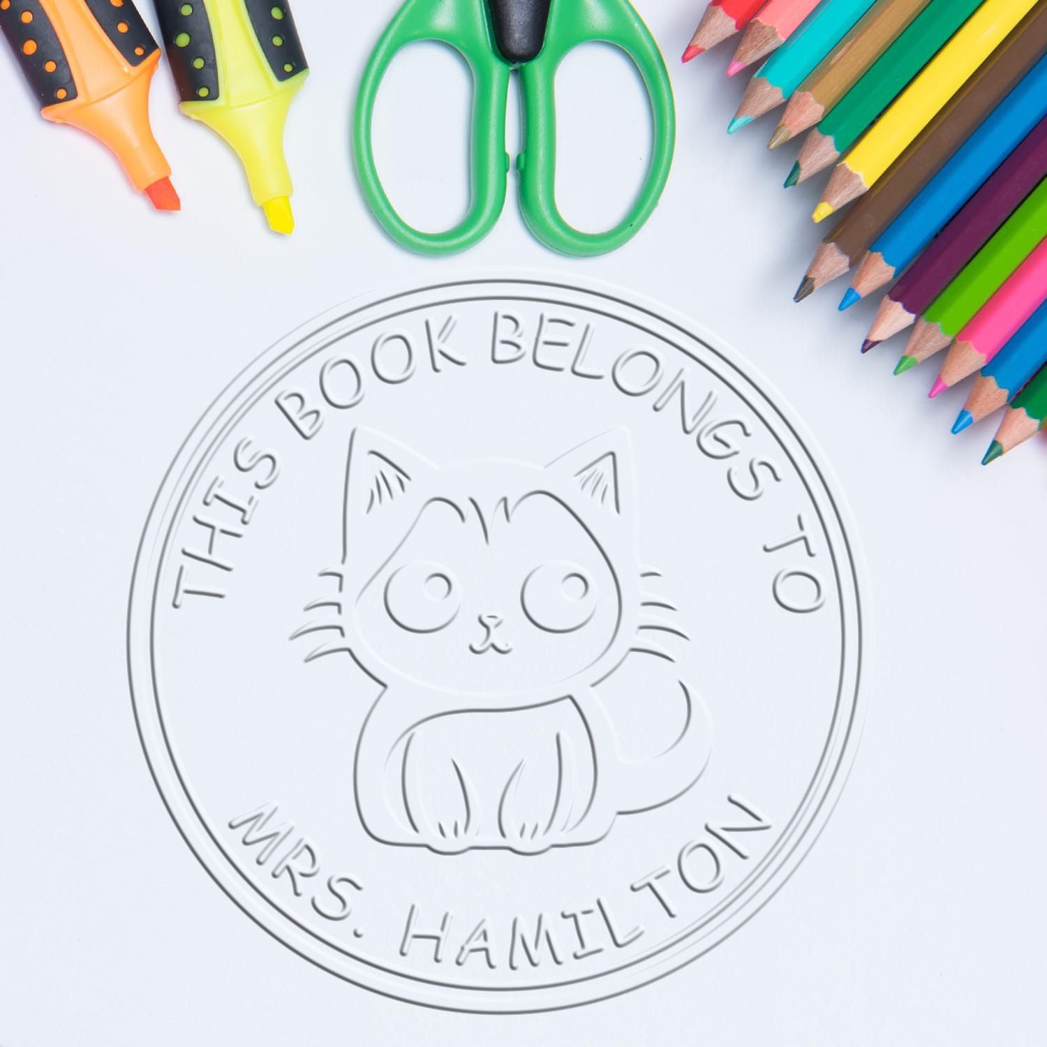 Round embossed cat book stamp and seal reading "This Book Belongs to Mrs. Hamilton," with colorful pencils, scissors, and highlighters.