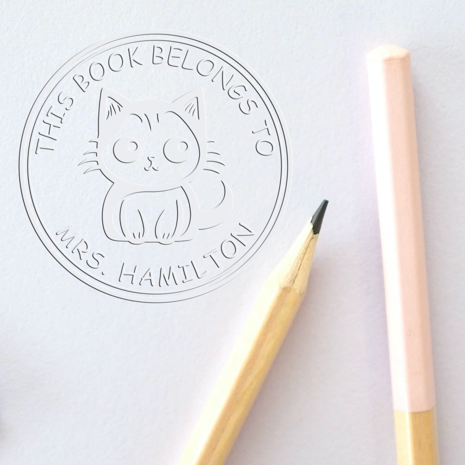 Embossed cat book stamp seal in pale gray on white paper, beside pencils, with This Book Belongs to Mrs. Hamilton text.