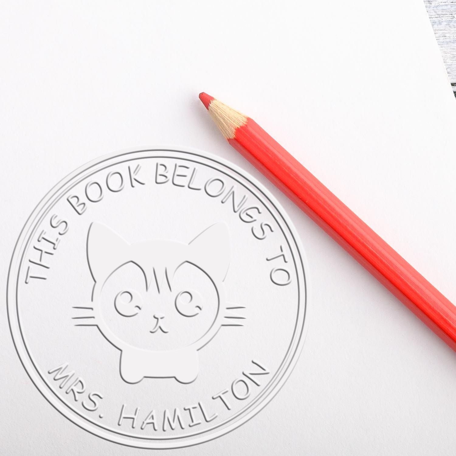 Embossed gray custom book stamp seal with cat outline beside a red pencil on white paper