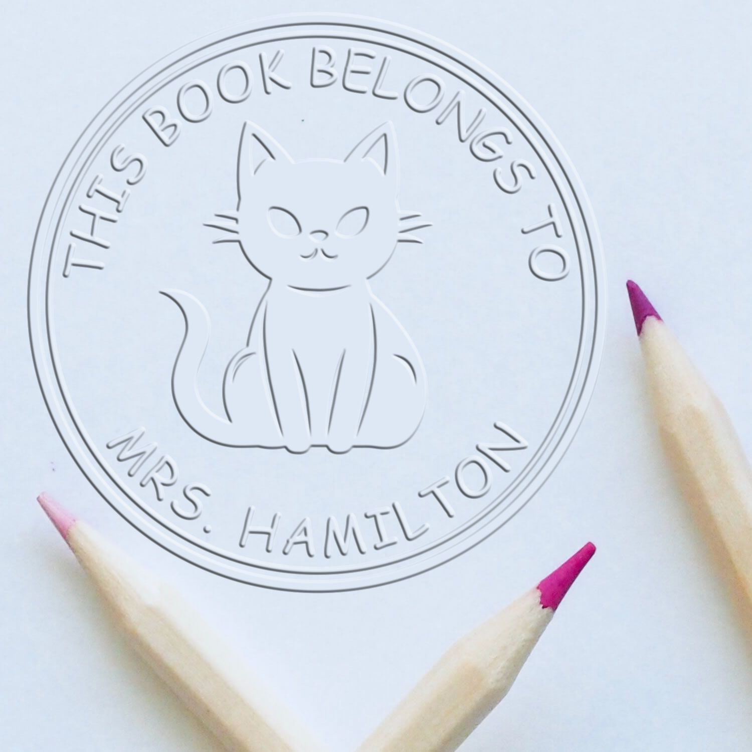 Embossed white book-ownership stamp seal with cat design and circular text, accented by pink pencil tips on a pale background.