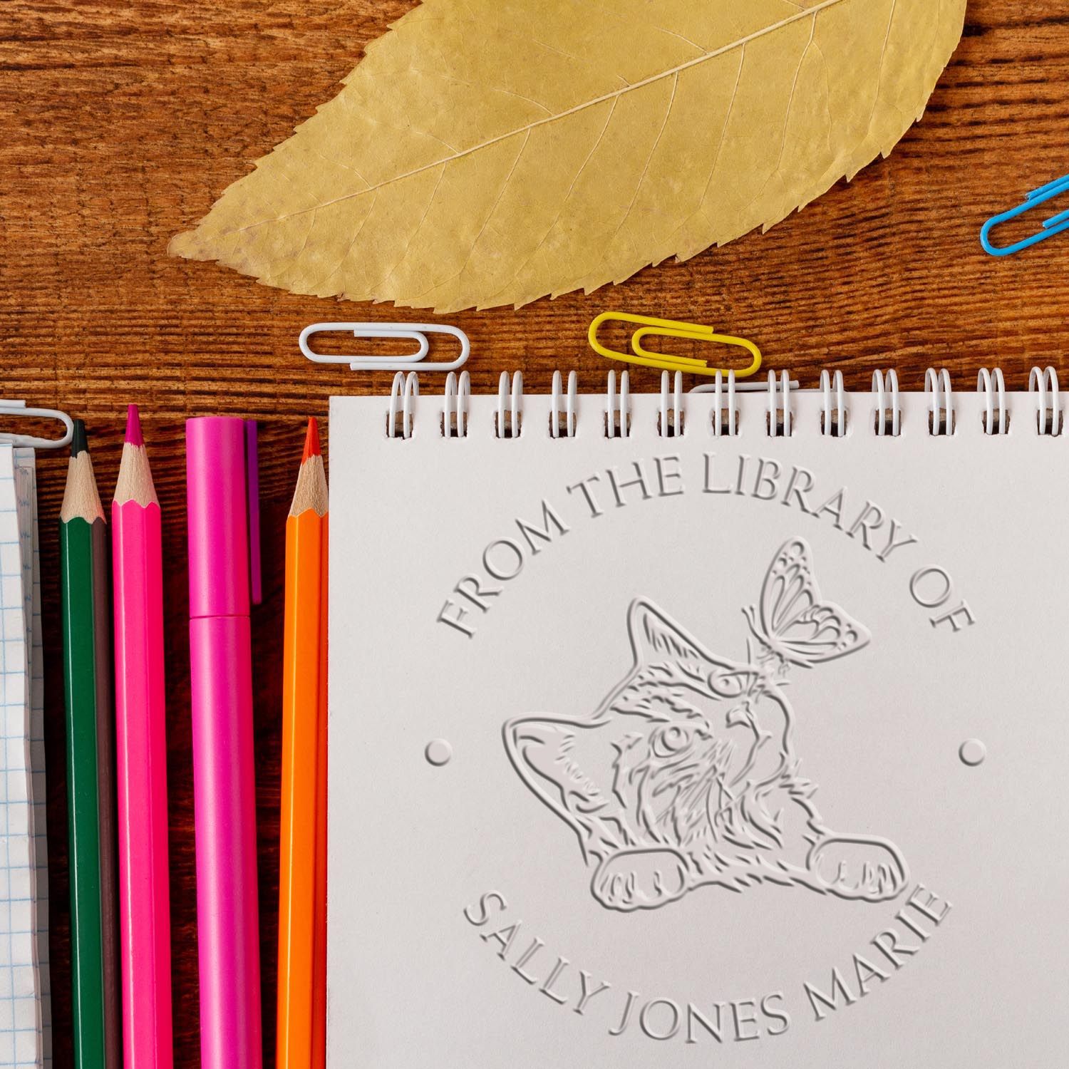 Embossed library seal notebook with cat and butterfly, beside colorful pencils, paper clips, and leaf on wood desk.