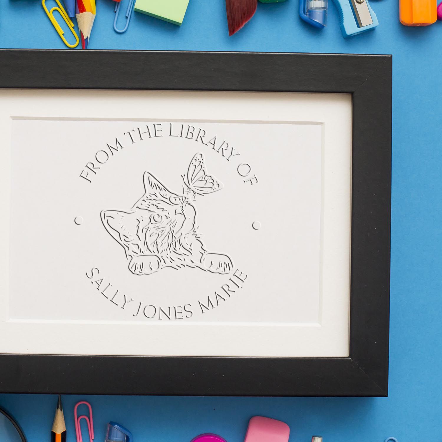 Black framed white embossing stamp seal with cat and butterfly, custom name design on blue background.