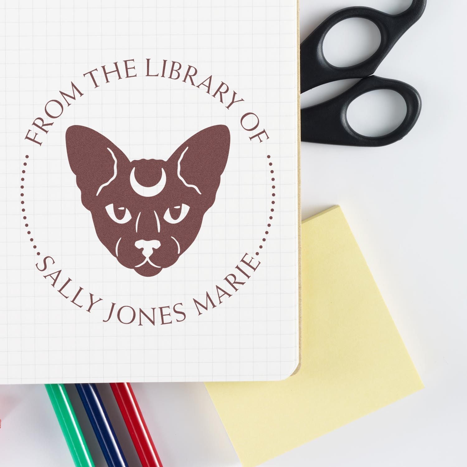 Brown custom library stamp seal with cat face and circular text, beside black scissors, colored pens, and a yellow sticky note.