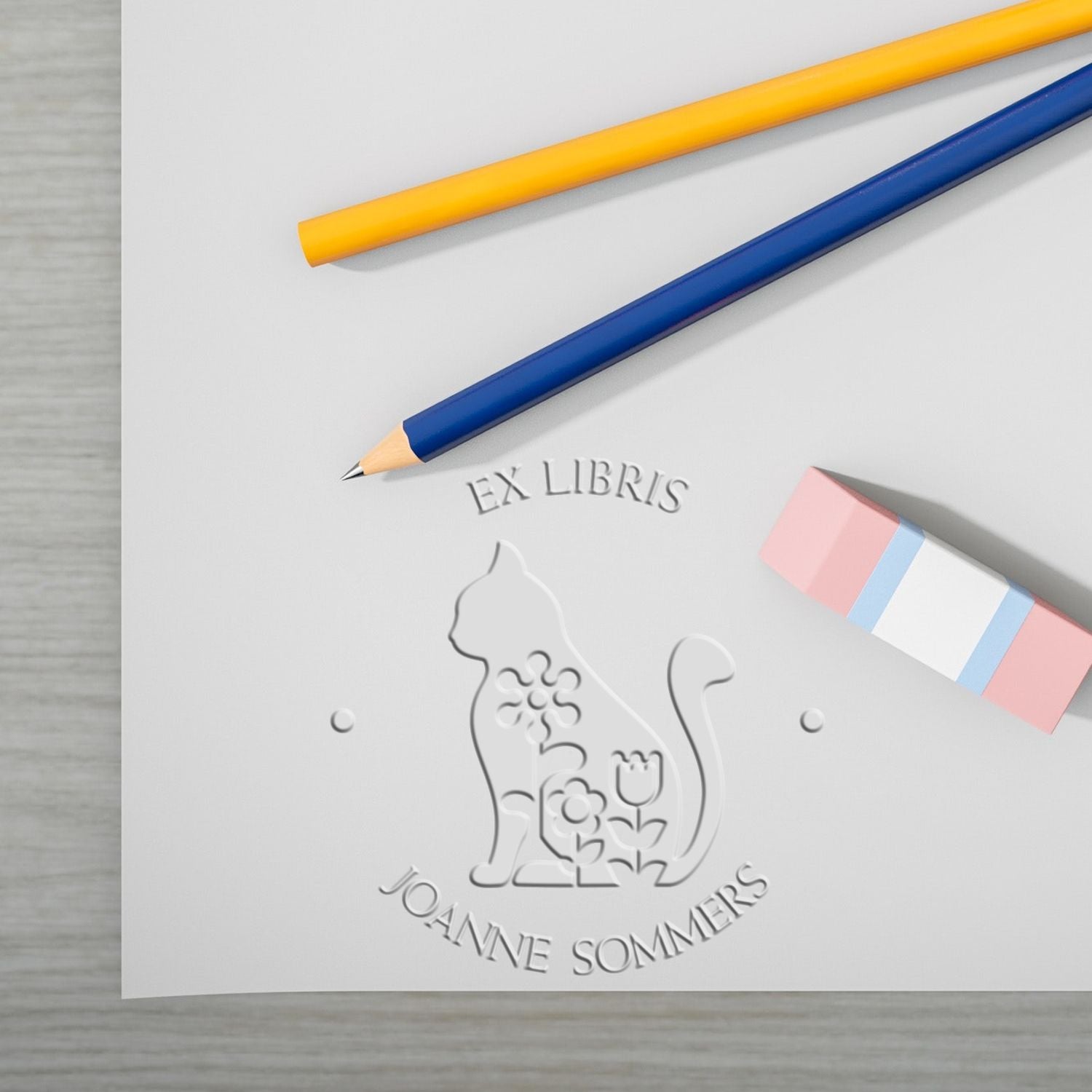 Yellow and blue pencils with pink eraser beside embossed cat ex libris book stamp and seal on white paper.