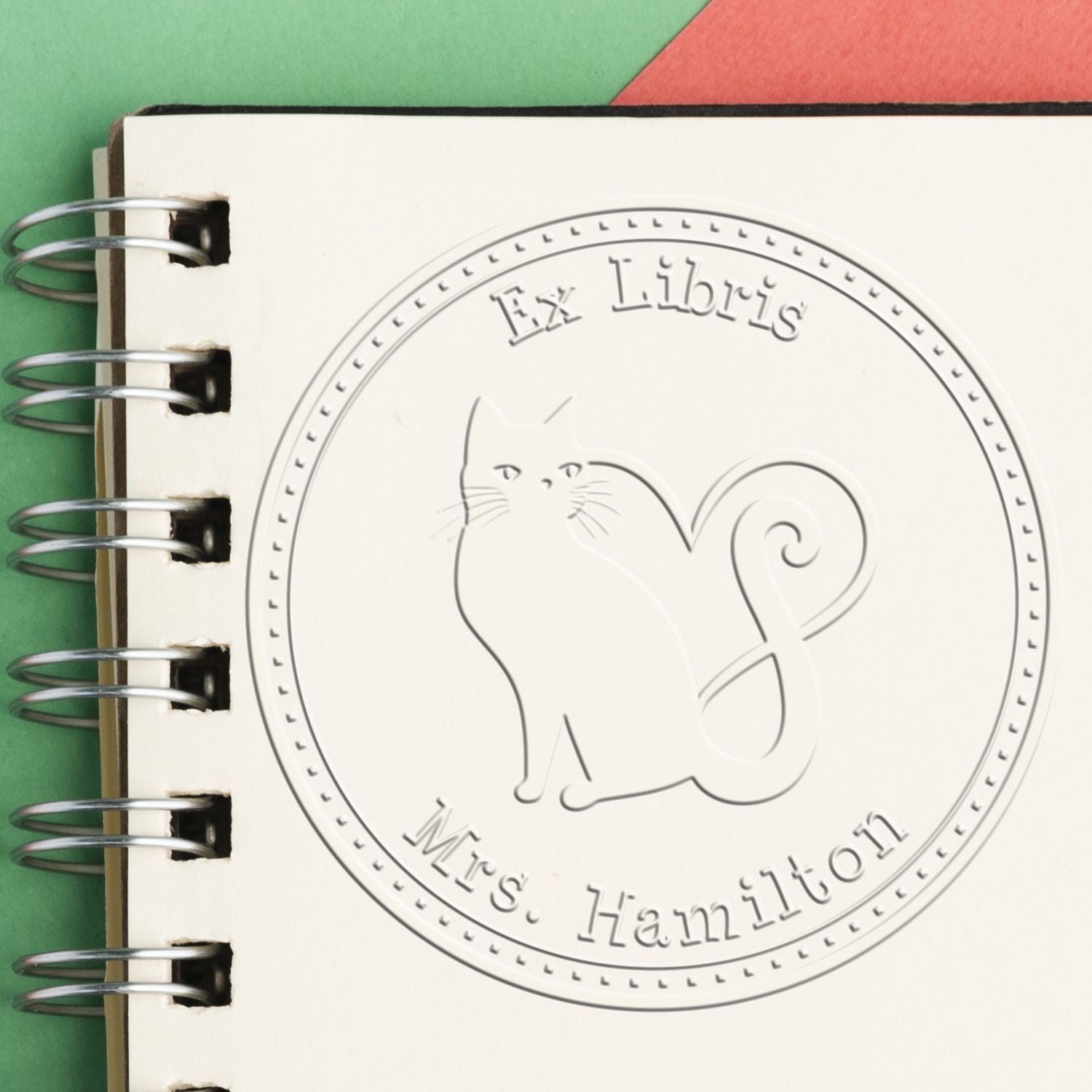 Embossed Ex Libris stamp seal with cat on cream spiral notebook, silver rings, against green and red background.