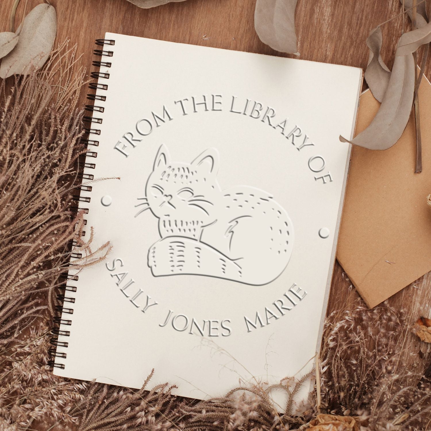 Cream spiral notebook with embossed cat stamp seal, reading From the Library of Sally Jones Marie, on a warm brown rustic backdrop.
