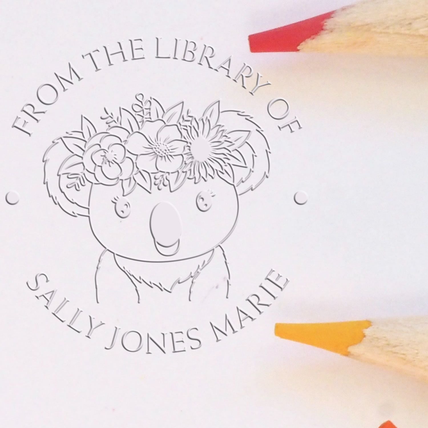 Personalized library stamp and seal with embossed gray koala and flower crown, circular text, on white paper beside red and yellow pencils.