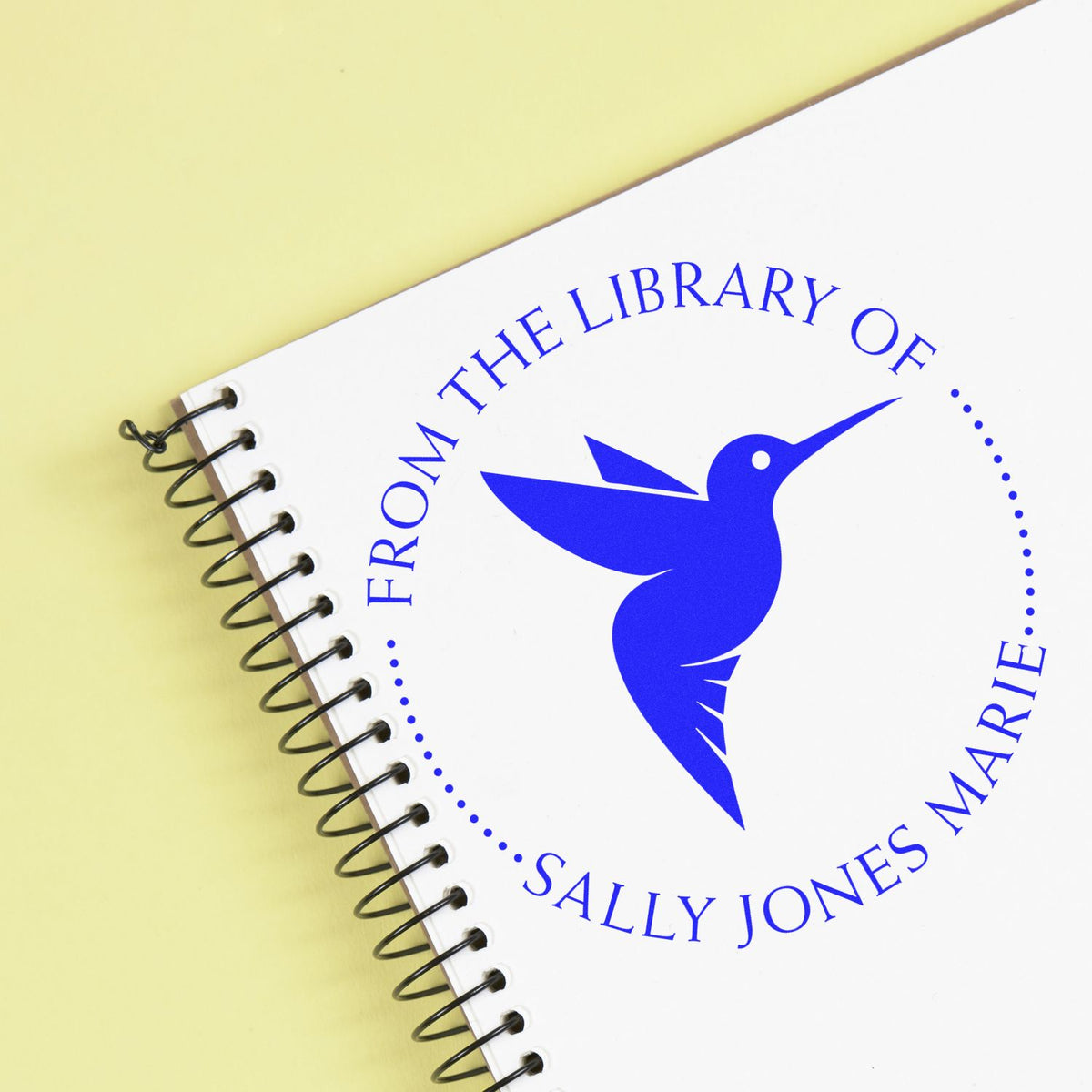 Blue custom library stamp/seal with hummingbird logo on white spiral notebook, on pale yellow background.
