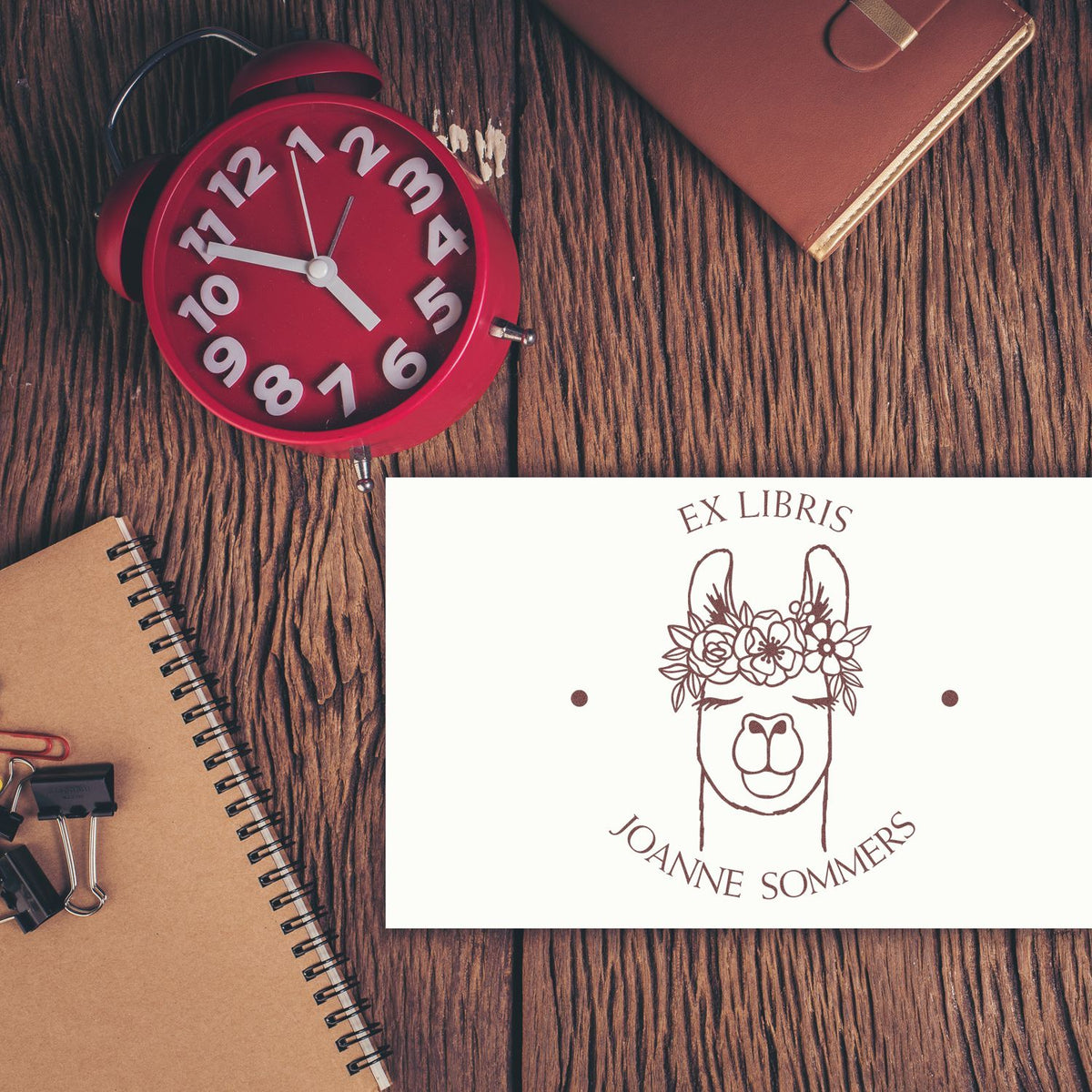 Custom ex libris stamp and seal with llama floral design on white card, beside a red alarm clock on a rustic wood desk.