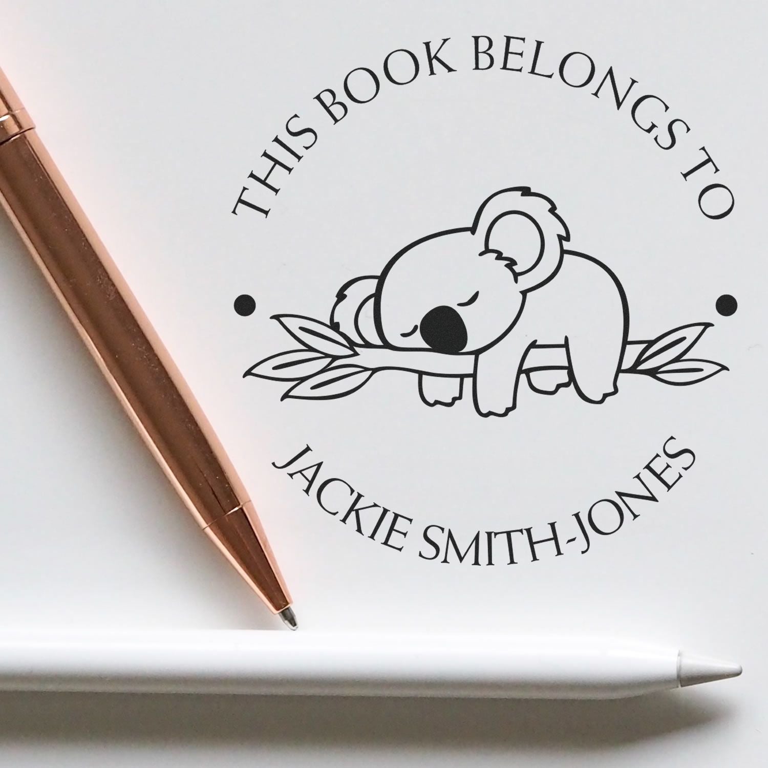 Personalized book stamp and seal reading This Book Belongs to Jackie Smith-Jones with sleeping koala, black text, rose-gold pen.