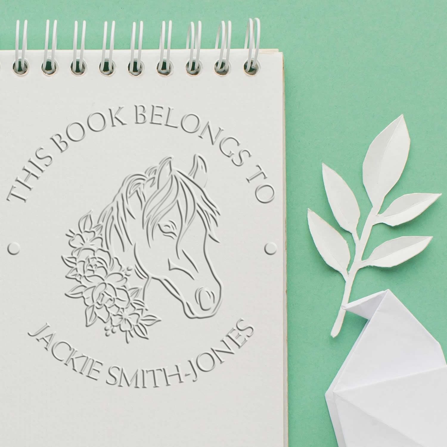 White embossed book stamp seal with horse and floral design on a notebook, on a mint green background with paper leaves.