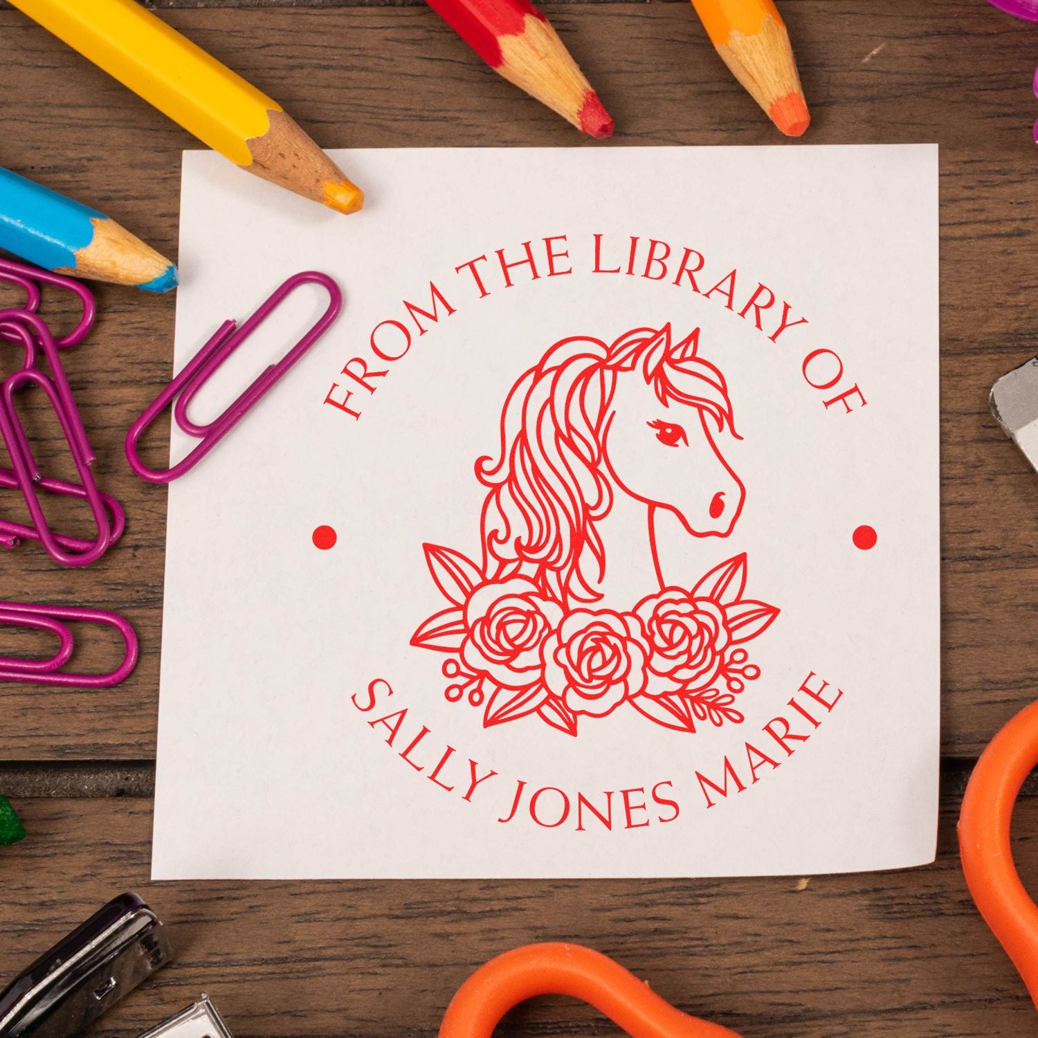 Custom red library stamp seal with unicorn and flowers on white card, surrounded by colorful pencils and purple paper clips