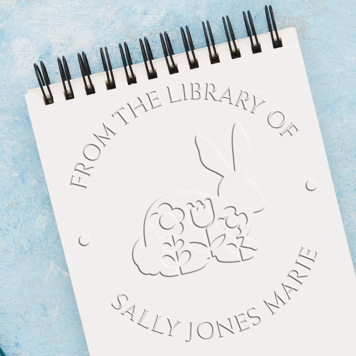 White spiral-bound embossing seal sample with raised rabbit and flowers, gray lettering, on a light blue background.