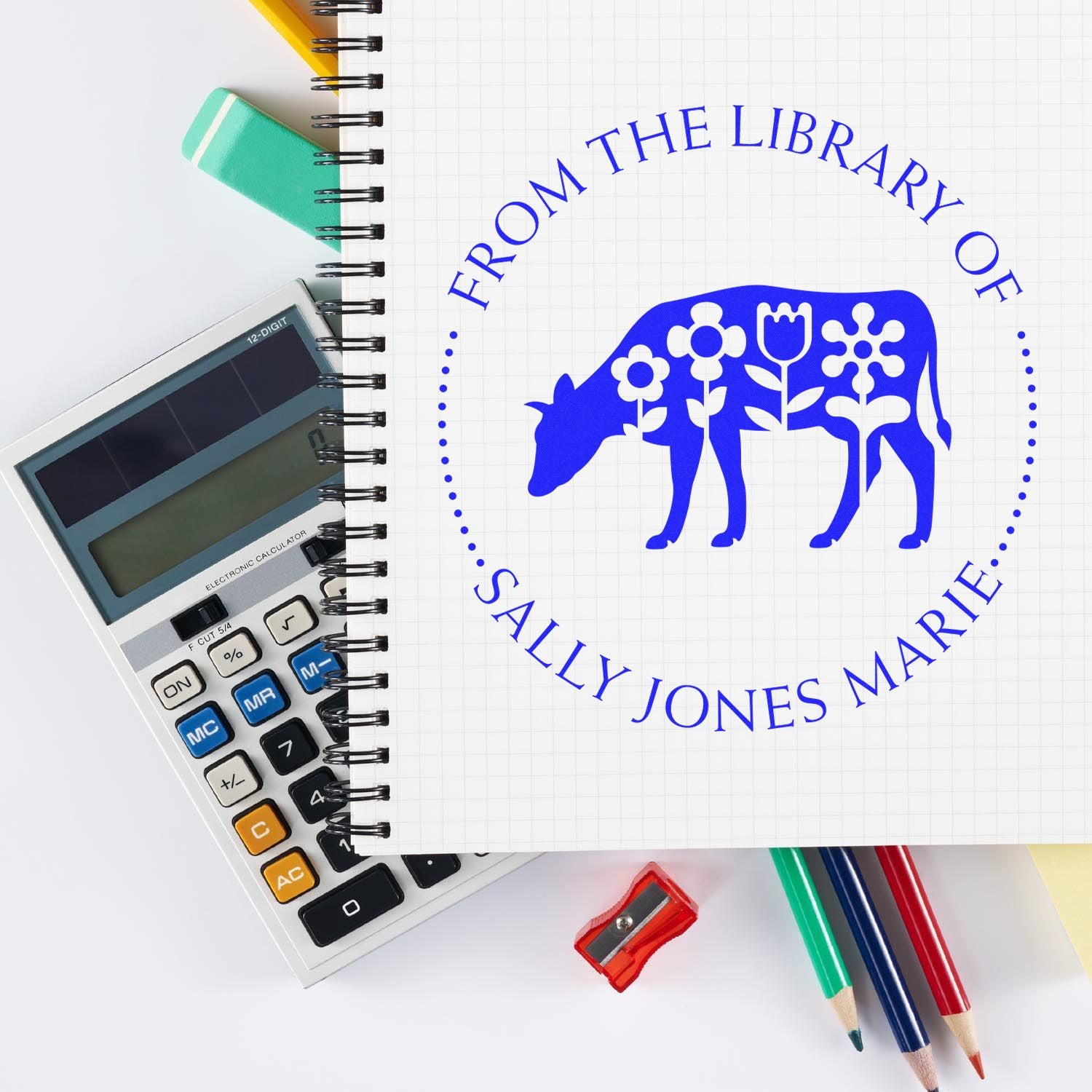 Blue custom library stamp seal with floral cow design on notebook, beside calculator, pencils, eraser and sharpener.