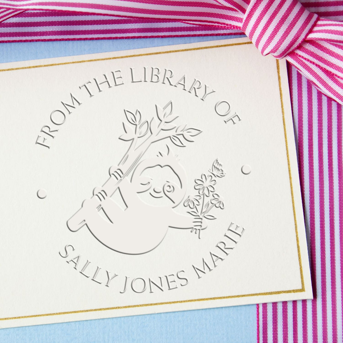 Custom embossed library seal on white card with sloth and flowers, gold border, pink-striped ribbon on blue background.