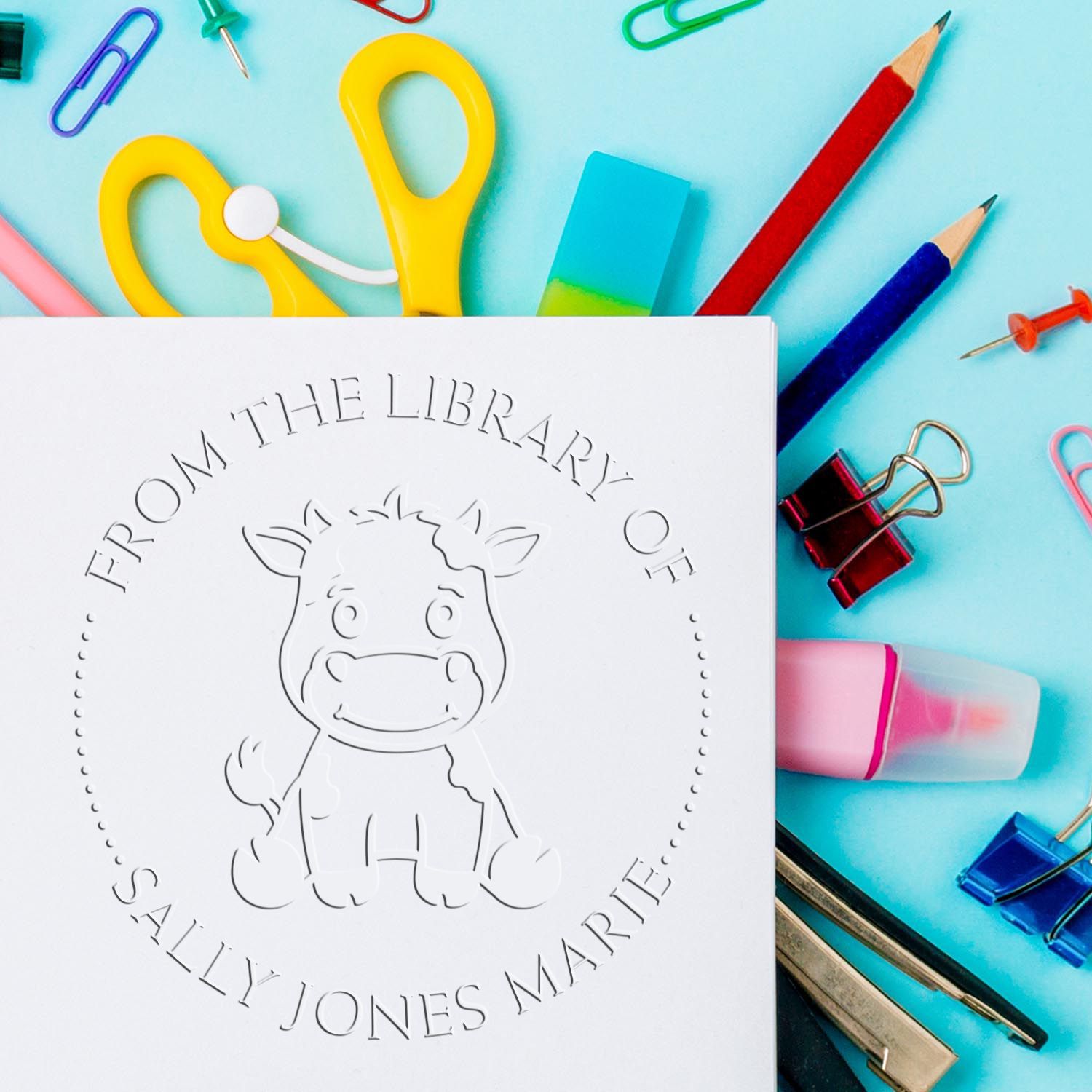 White embossed stamp seal with baby hippo and circular text, surrounded by colorful stationery on a turquoise background.