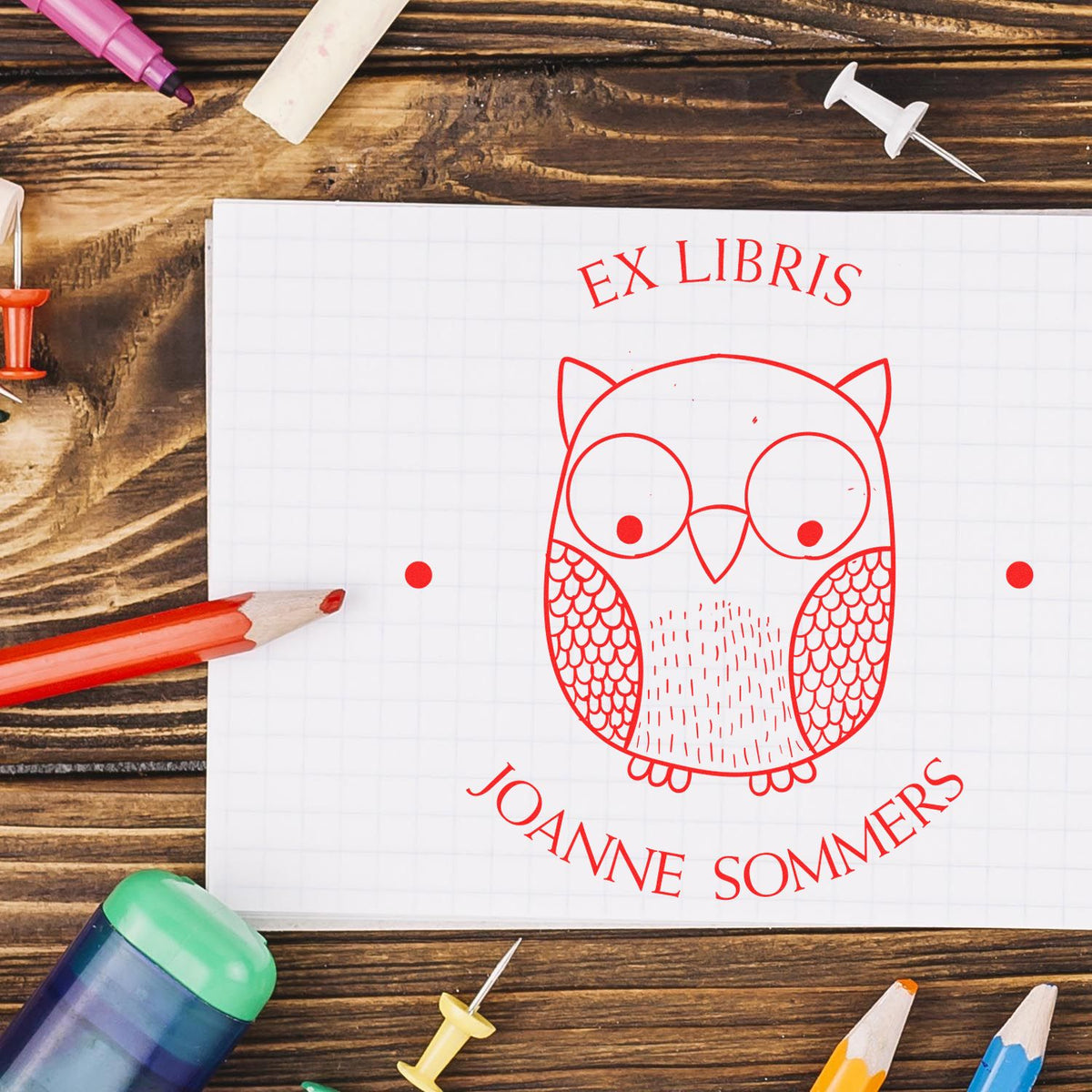 Red owl ex libris custom stamp and seal on white grid paper, with colored pencils on a rustic wood desk.