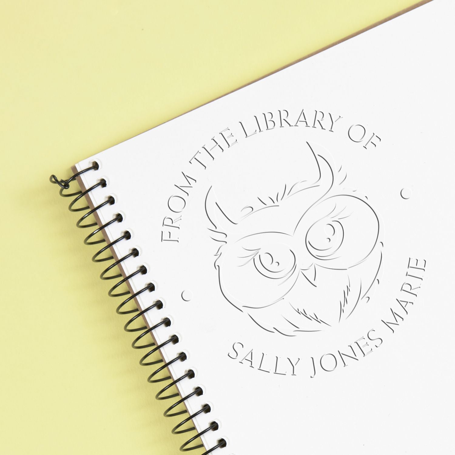 White spiral-bound notebook with embossed owl library stamp seal, black coil, on pale yellow background.