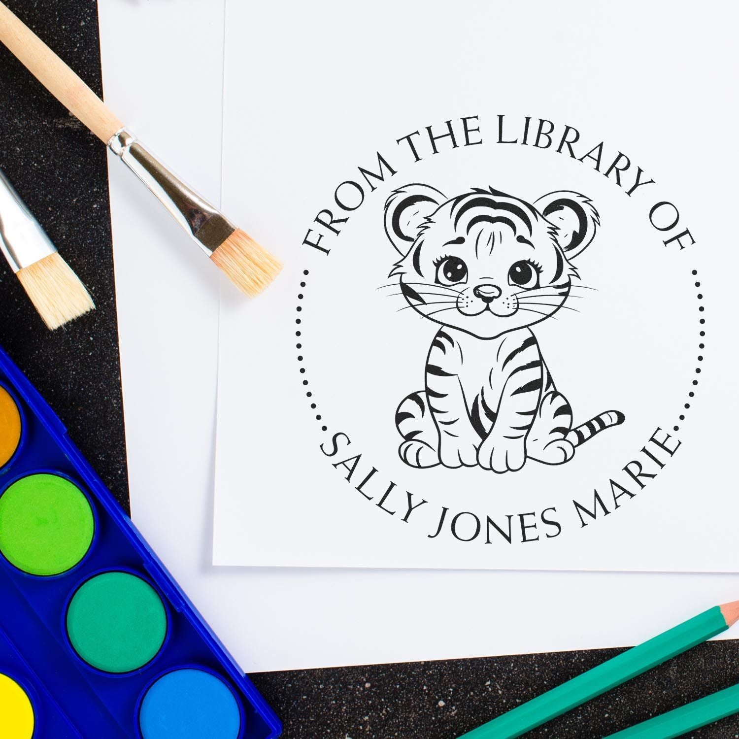 Black custom library stamp seal with tiger cub and Sally Jones Marie on white paper, beside paintbrushes and bright watercolor paints.