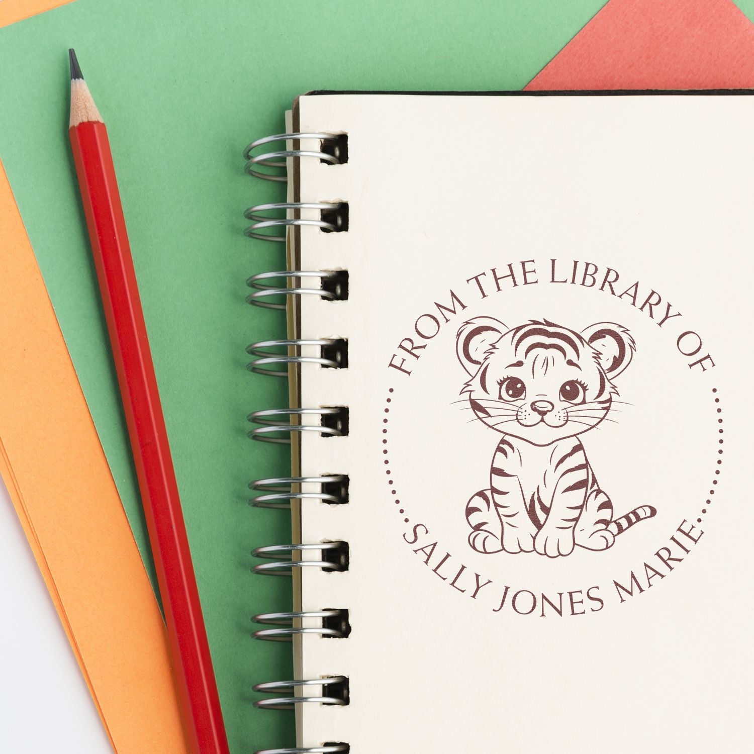 Cream spiral notebook with brown tiger library stamp and seal, beside a red pencil on green, orange, and coral paper.