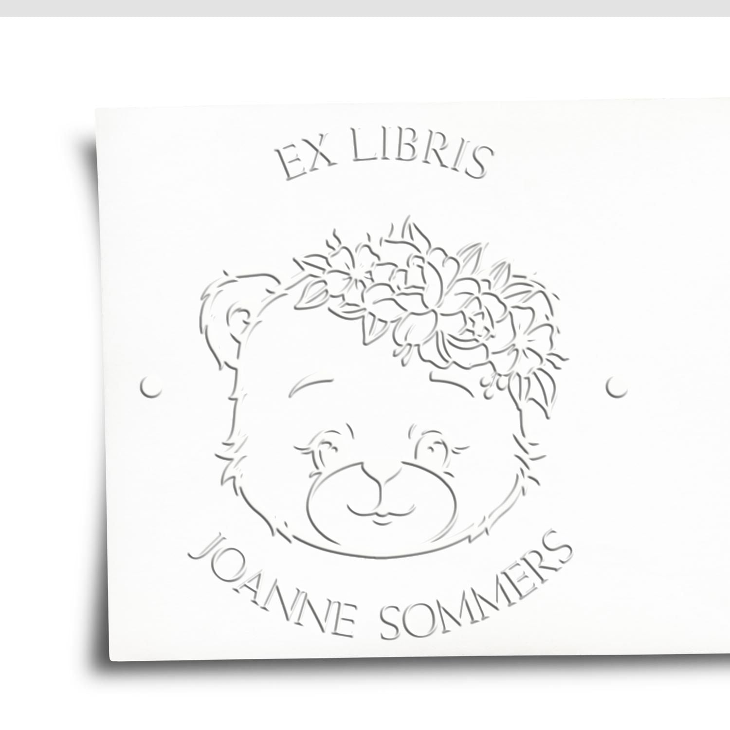 White embossed ex libris bookplate seal with a bear wearing a floral crown and the name Joanne Sommers.