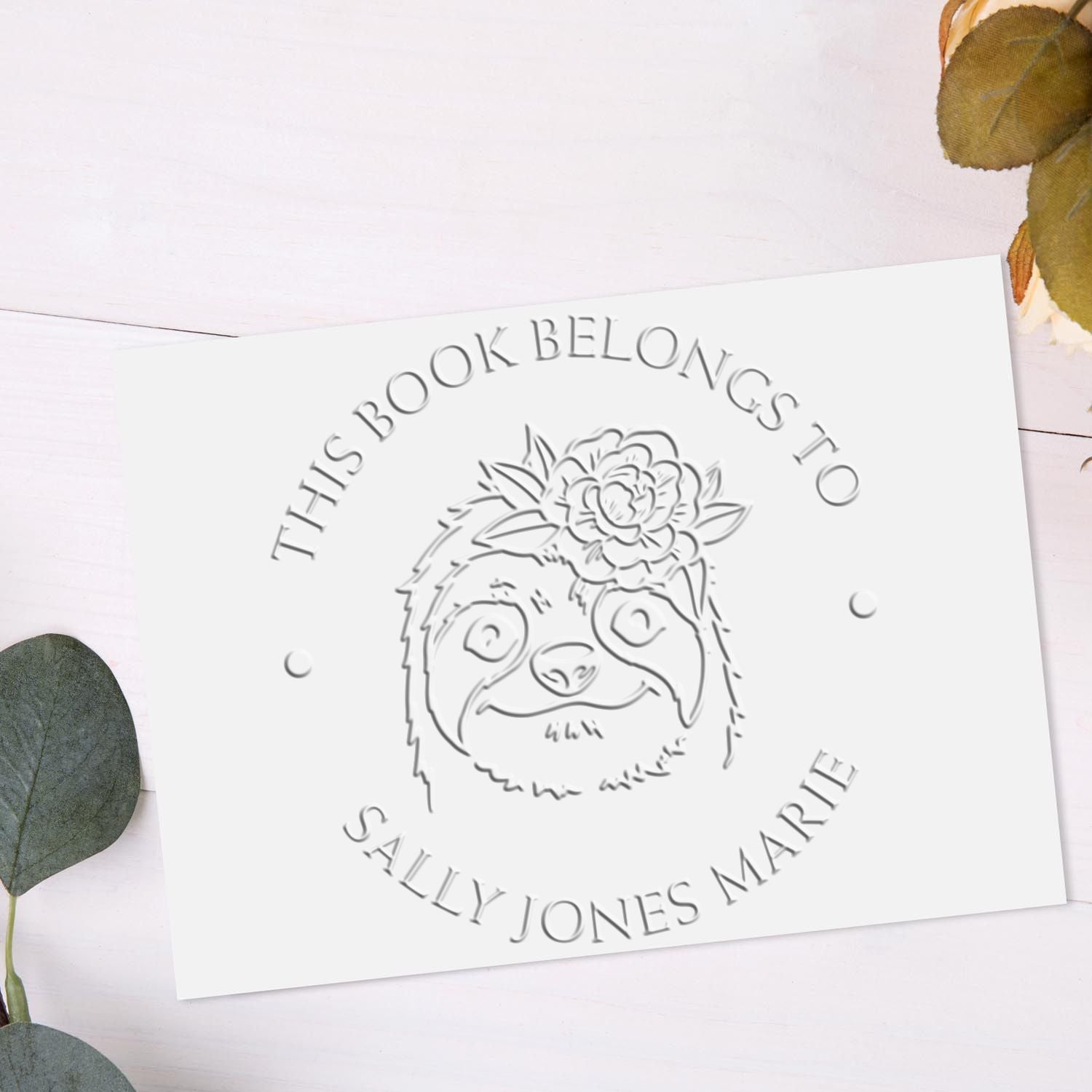 Custom embossing seal stamp with sloth outline and Sally Jones Marie text on white card, accented by eucalyptus leaves
