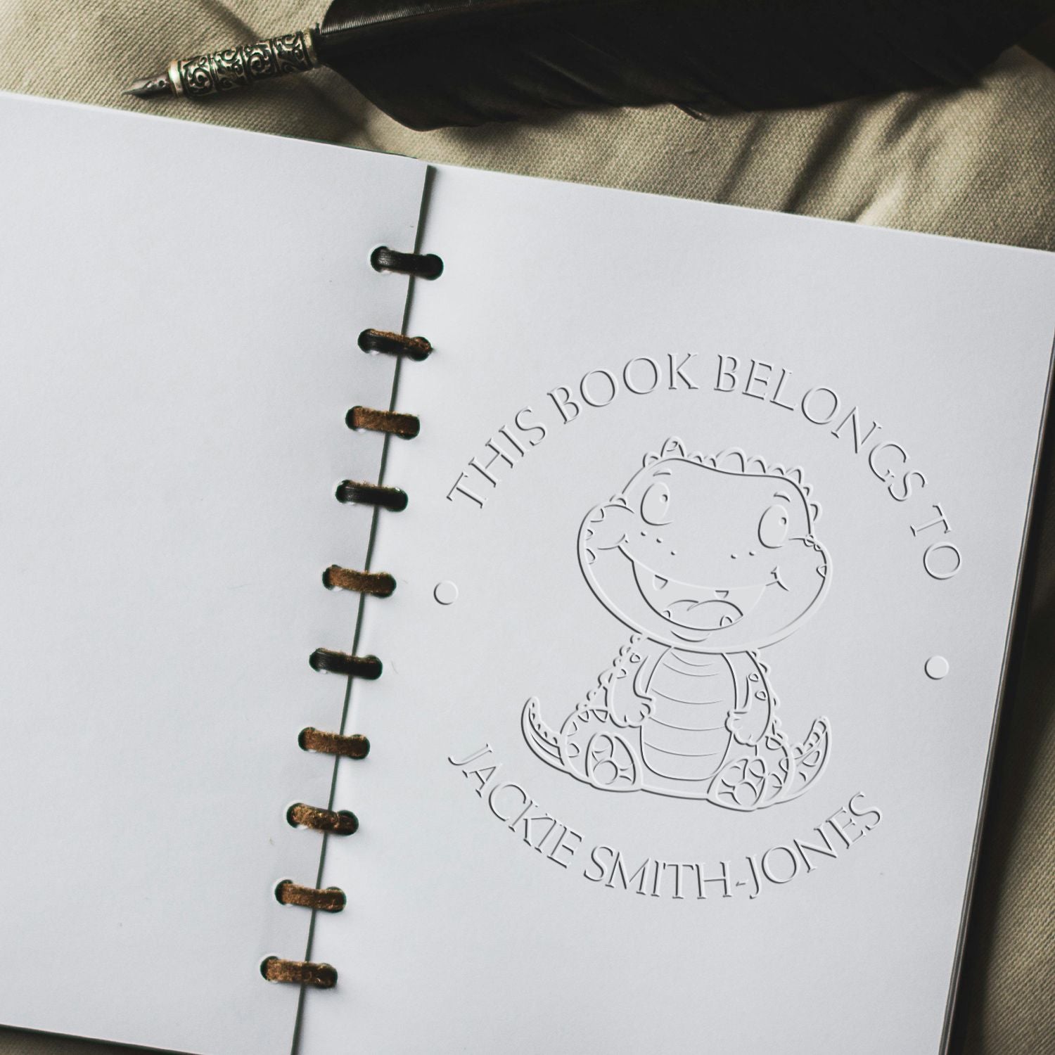 Embossed white book seal stamp with dinosaur and This Book Belongs to Jackie Smith-Jones on spiral-bound pages.