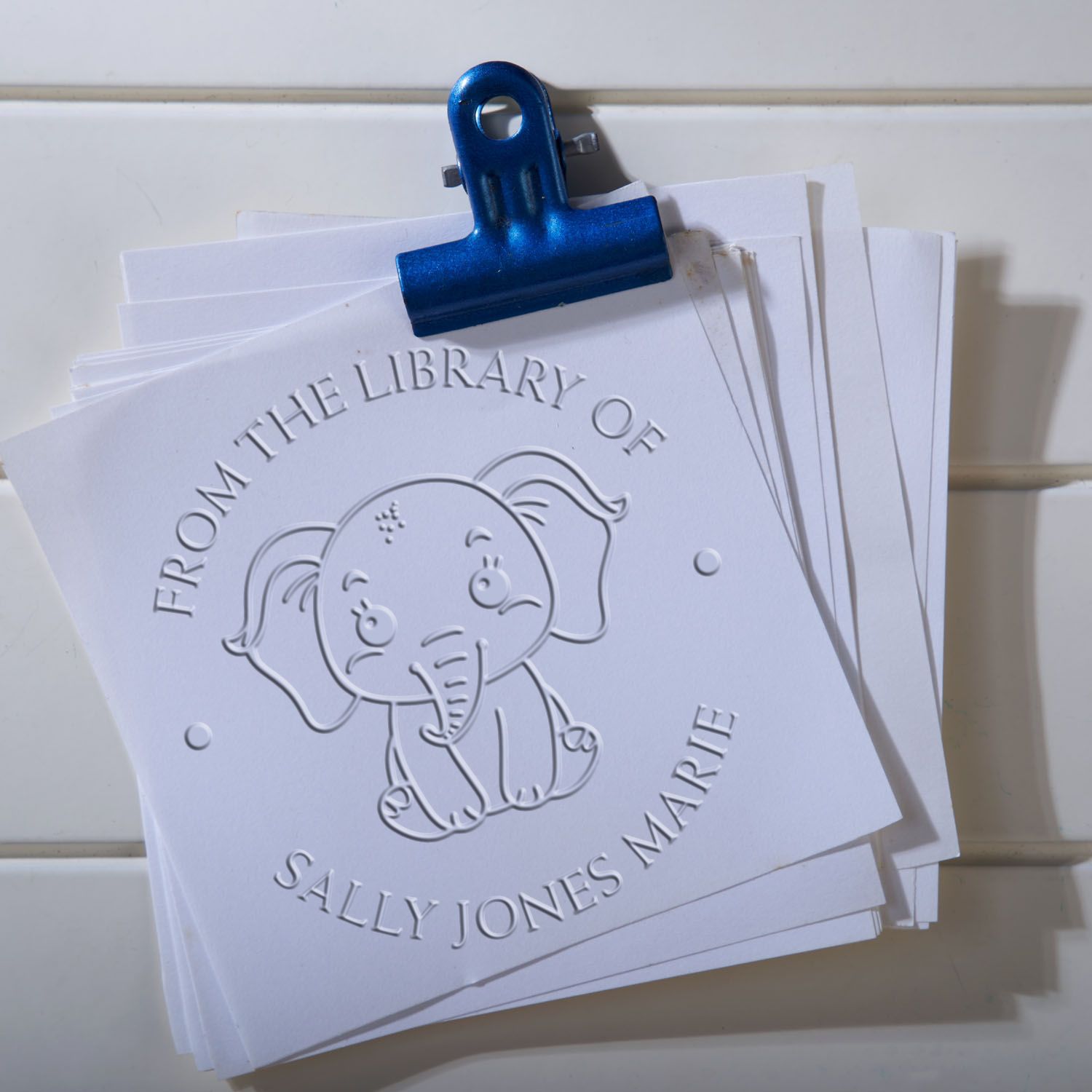 Blue binder clip holding white embossed library stamp seals with elephant design and From the Library of Sally Jones Marie text.