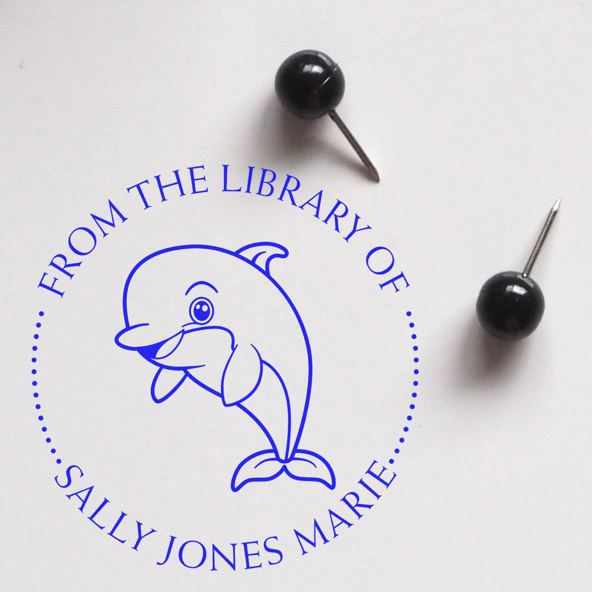 Blue custom library stamp and seal with dolphin logo and circular text, beside two black push pins on white background.