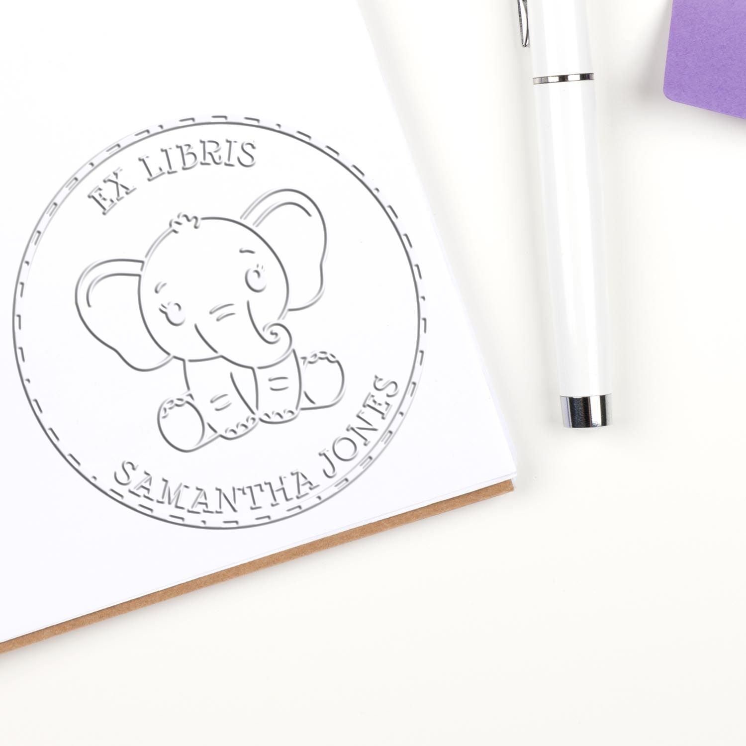 Custom white embossing seal stamp with elephant logo and family name, beside a white pen and purple notebook on a white desk.