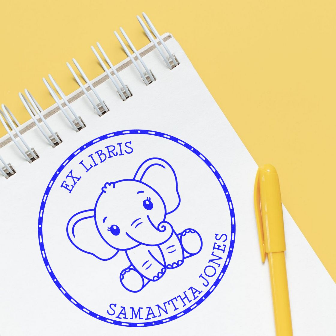 Blue custom elephant stamp seal on a white spiral notebook with a yellow pen and yellow background.
