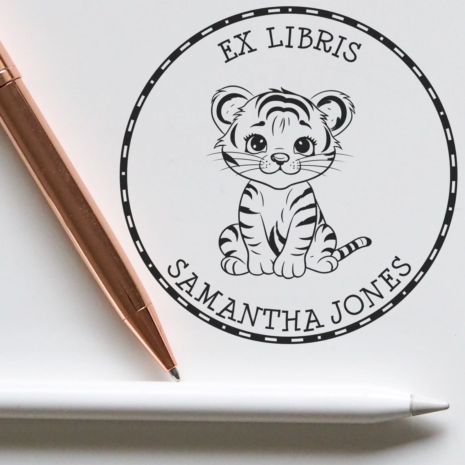 Black custom ex libris stamp seal with cute tiger cub and Samantha Jones text, shown with rose gold pen and white stylus.