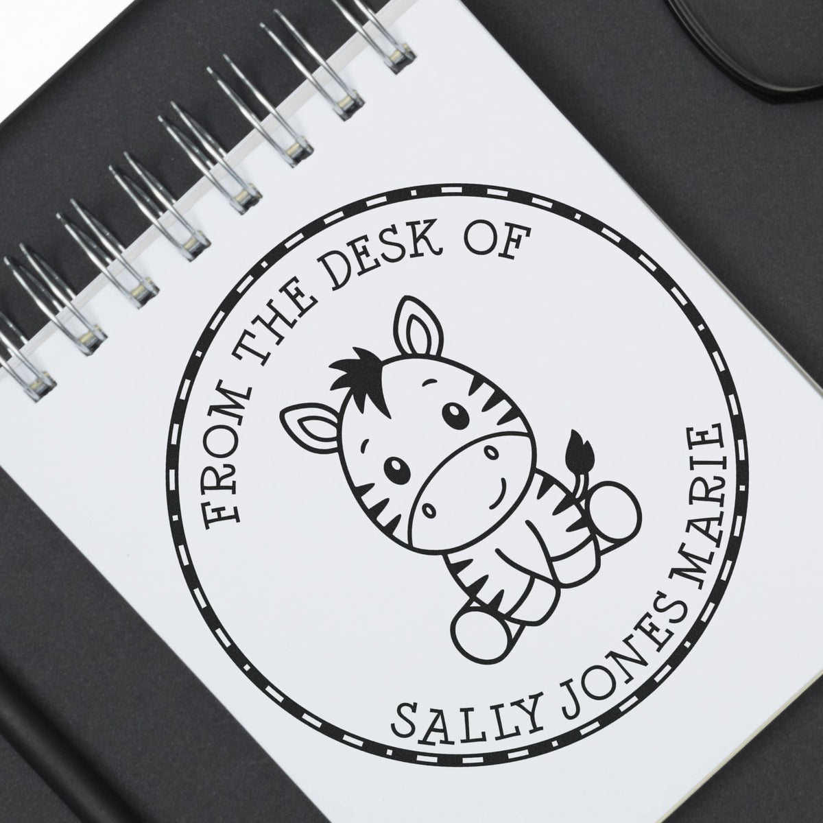 Black circular stamp seal with zebra mascot and From the Desk of Sally Jones on a white spiral notebook.