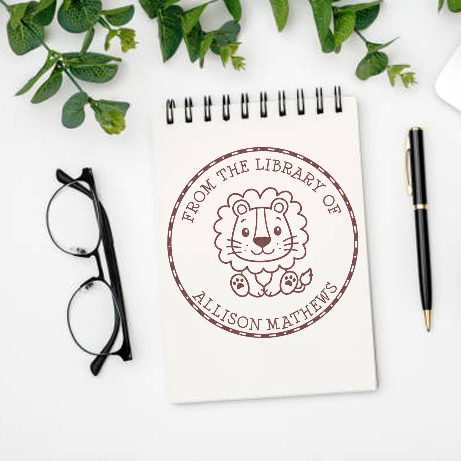 Brown library stamp seal with lion illustration and Allison Matthews text on a white spiral notebook, with glasses, pen, and green leaves.