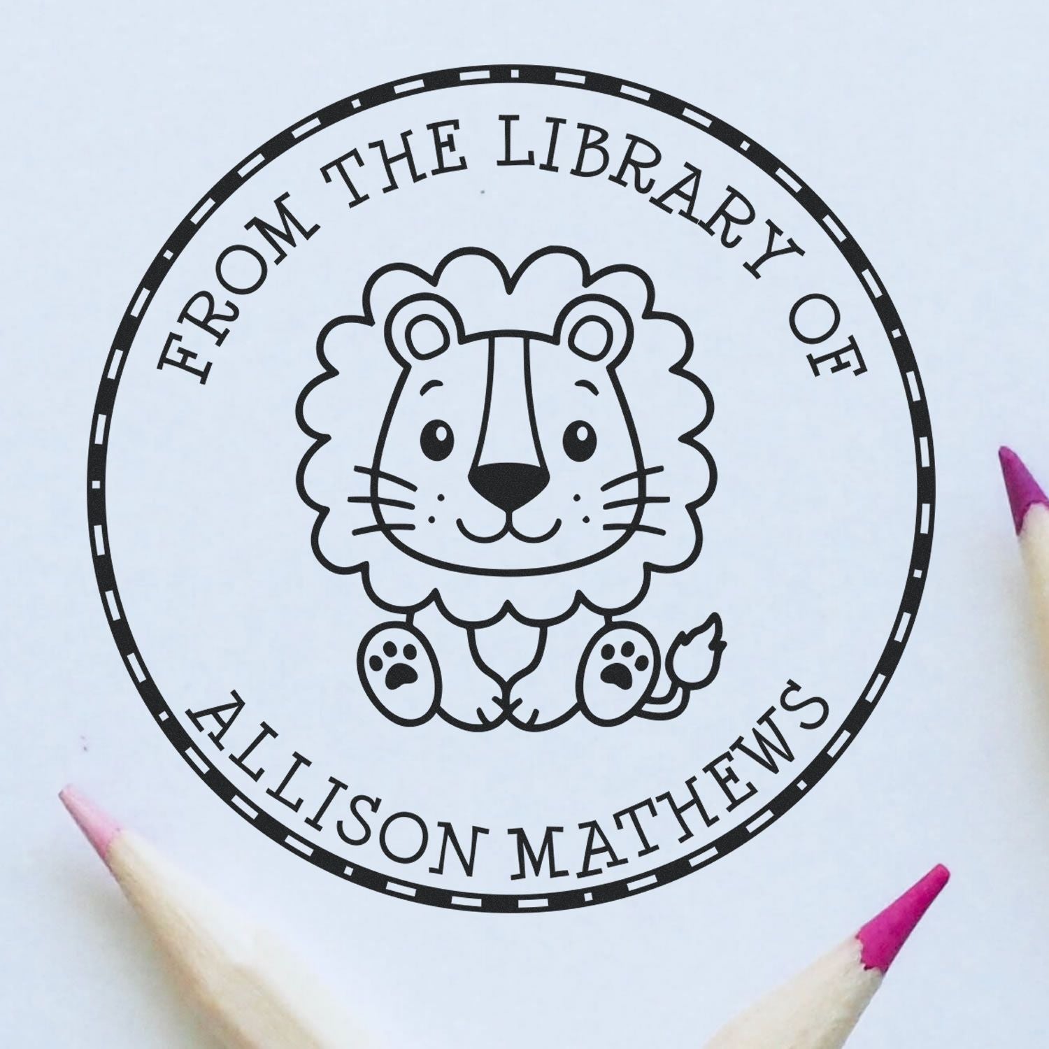 Black circular library stamp seal with lion logo and From the Library of Allison Mathews on white paper, pink pencils nearby.